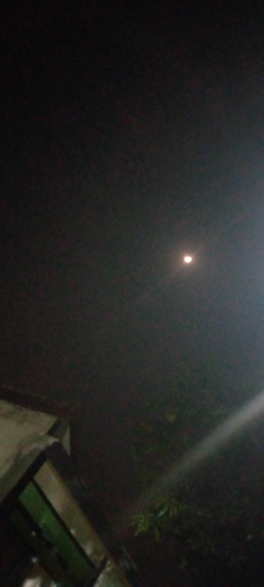 A nighttime shot of a waning crescent moon in a dark, hazy sky over Sumberwono, Indonesia. A sliver of a building with green-tinted windows is visible in the lower left corner, angled upwards. The scene appears to be outdoors, with hints of foliage and a bright light source, possibly a streetlamp or a high-intensity light, casting a beam across the lower right portion of the frame. The image is blurry and dark, with the moon being the most distinct subject.