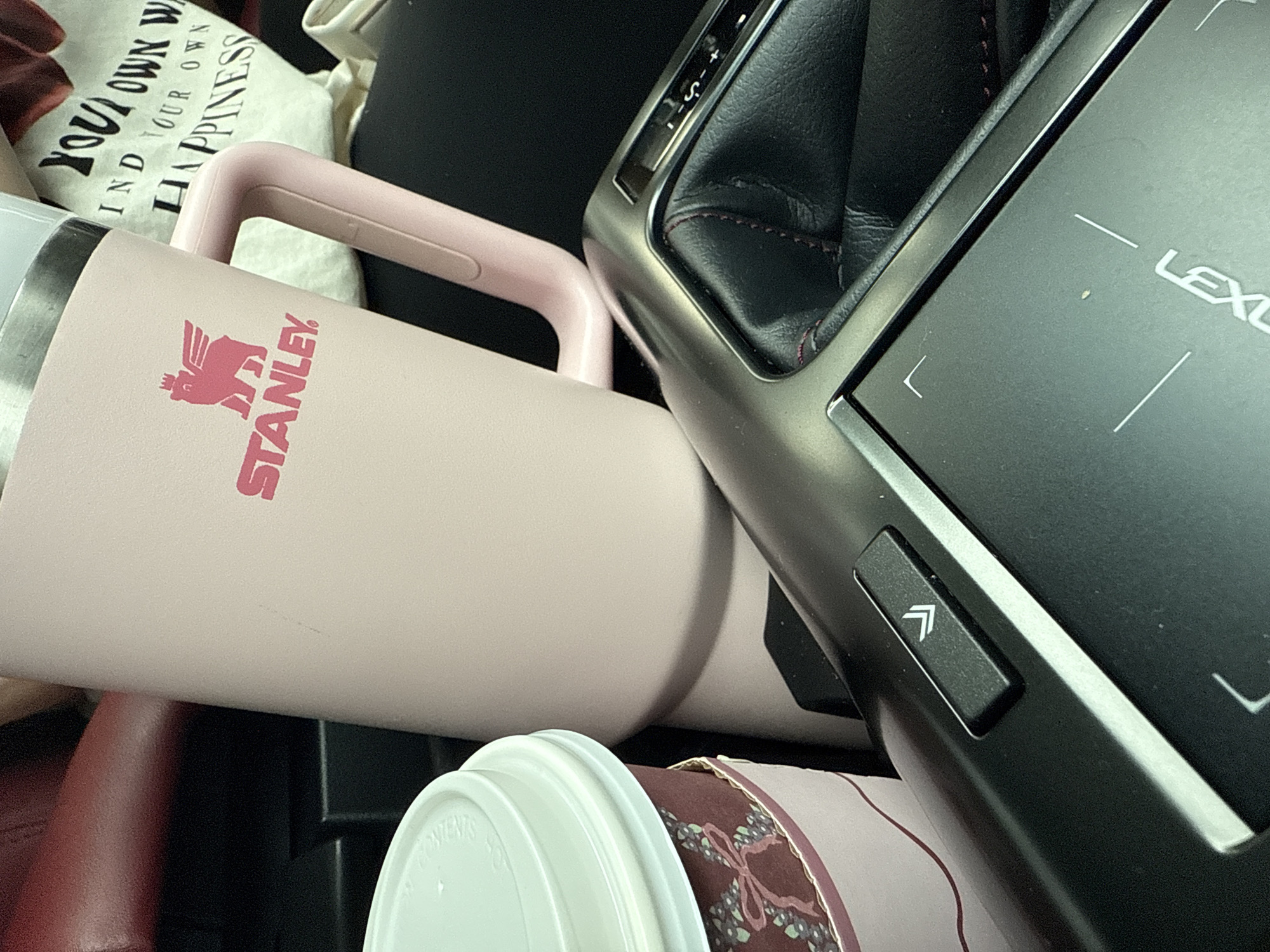 The image shows a close-up view of a car's center console, with various items scattered around. A pink Stanley mug with a handle is prominently displayed, obscuring part of a paper coffee cup with a white lid. The car's gear shift and infotainment system, labeled "LEXUS," are also visible. A reusable tote bag with the phrase "YOUR OWN W-IND YOUR OWN HAPPINESS" is partially visible in the background. The location is likely inside a car in Davie, United States of America, as indicated by the city information. No people are visible in the image, and no specific actions or interactions are captured. The environment is limited to the interior of the car.
