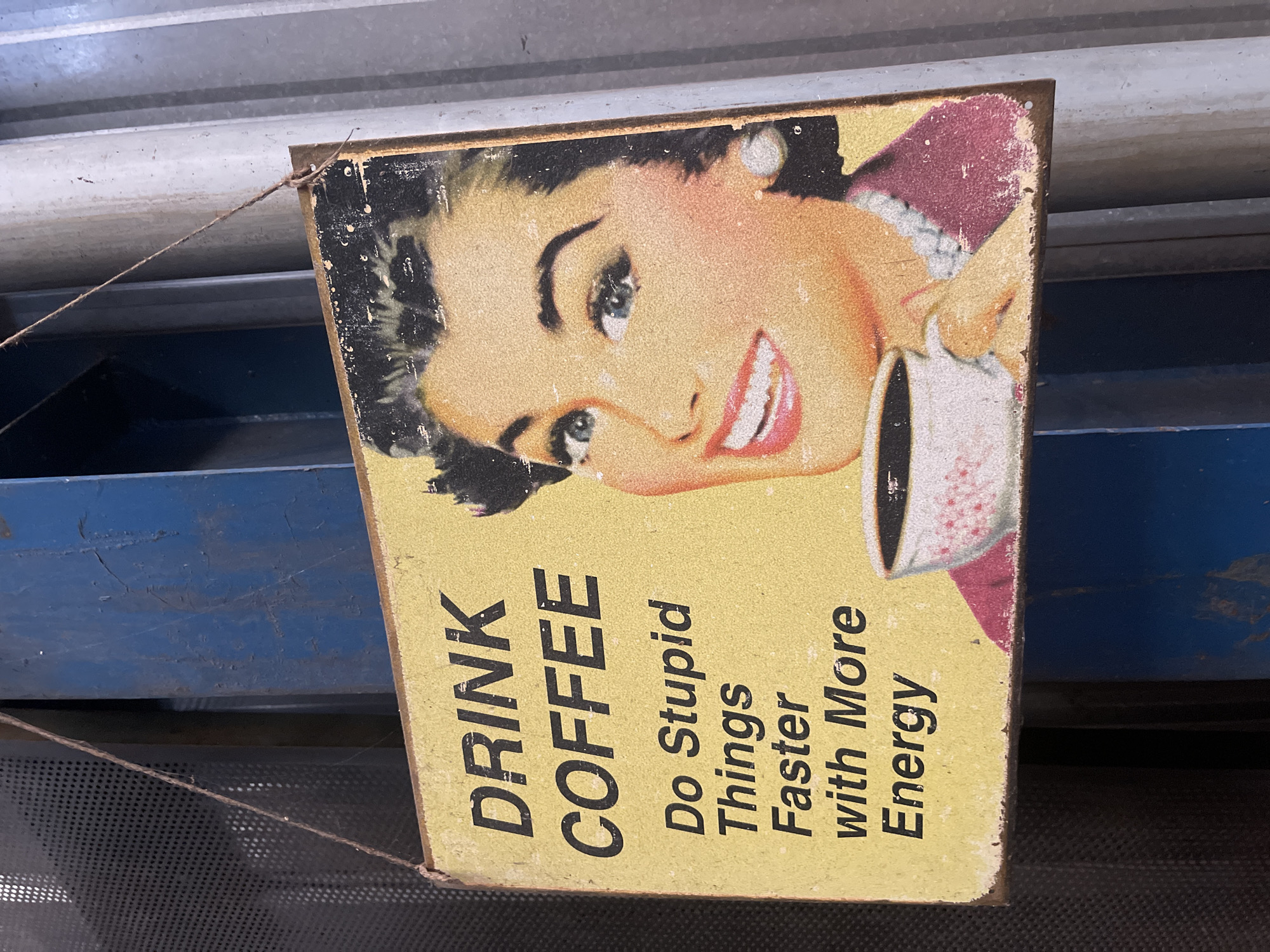 The image depicts a vintage-style sign hanging in an indoor setting. The sign features a cartoon image of a woman with a coffee cup and the text "DRINK COFFEE Do Stupid Things Faster with More Energy". The sign has a yellow background and black lettering, and it shows signs of wear and tear. It is suspended by a thin string. The background reveals industrial-like elements, including metal structures with a mix of blue and grey tones. The exact location in Da Lat, Vietnam is not discernible from the image.