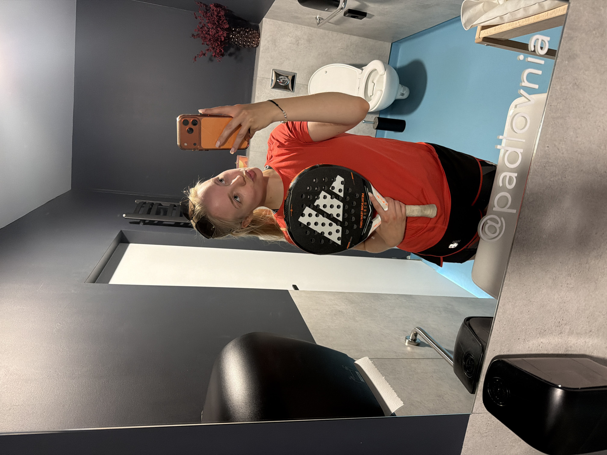 A mirror selfie taken in a restroom, location Unknown, Unknown. The subject is a blonde woman wearing a red shirt and black shorts, holding an orange smartphone in her right hand and a padel racket in her left hand. The mirror reflects a view of a toilet, a towel rack, and a flower decoration. In the foreground, there is a dark gray surface with a black dispenser on it. The mirror also has a watermark that says "@padlovnia".