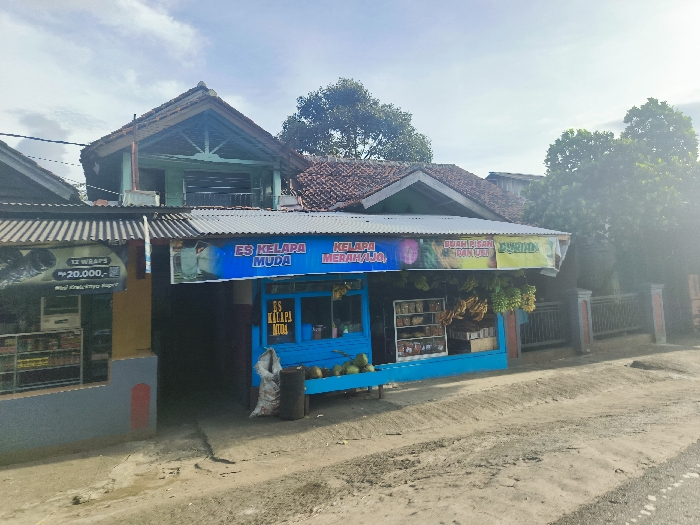 The image shows a storefront in Cikalong, Indonesia, at the coordinates -6.761 latitude and 107.447 longitude. The storefront has a blue awning with a sign that says "Es Kelapa Muda," which translates to "Young Coconut Ice." The storefront has a small window display and several shelves with various items for sale. There are several banana bunches hanging in front of the store. There is a bag of charcoal in the front. There is a building with a green roof to the left. In the background are several buildings and trees. The ground is made of gravel and dirt. The sky is clear and partly cloudy. The buildings are typical of the region, with corrugated metal roofs and simple construction. The trees are tropical and leafy. The scene is typical of a small town or village in Indonesia.
