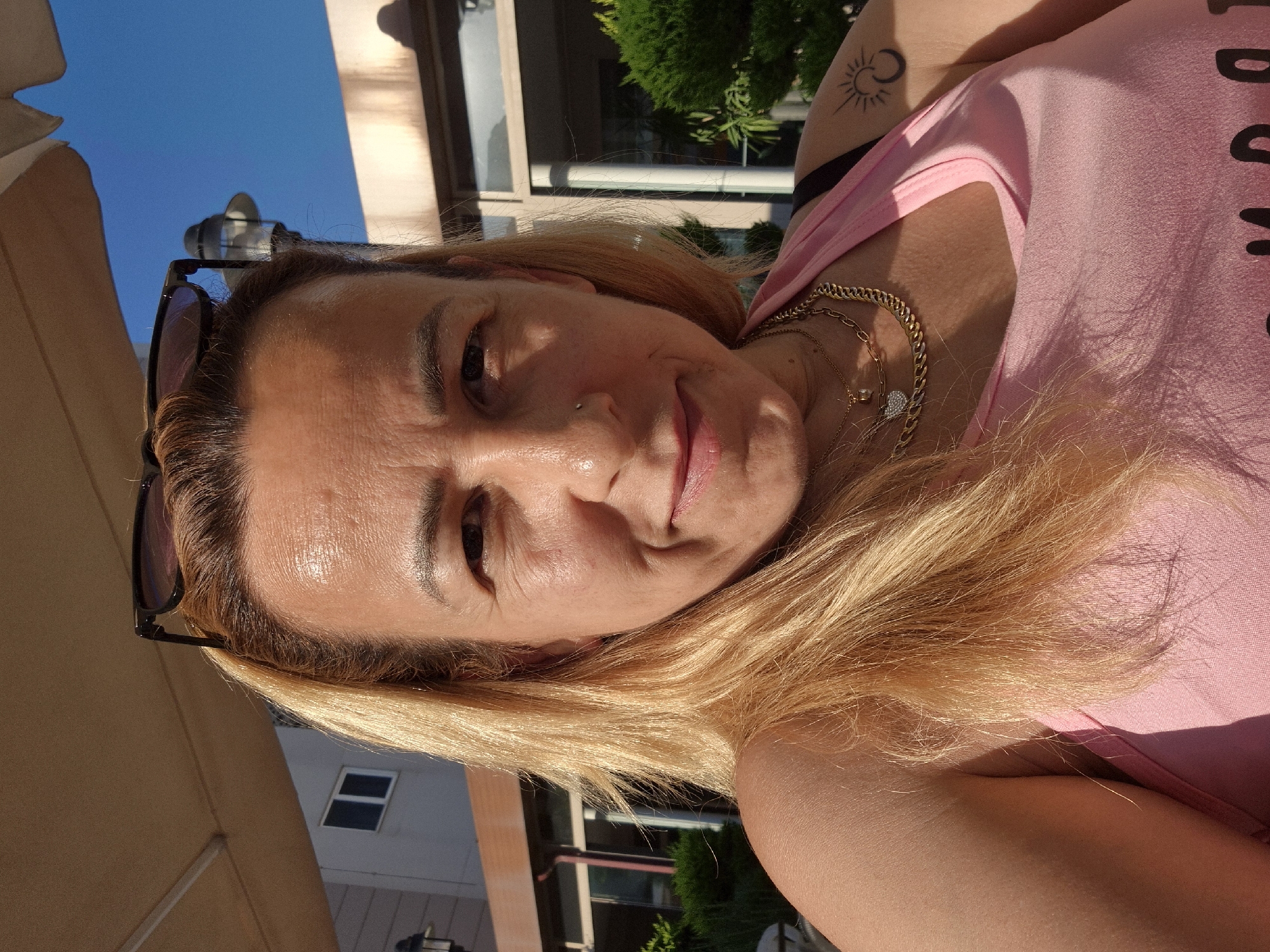 A head-and-shoulders portrait of a light-skinned woman with shoulder-length blonde hair is taken outdoors in daylight. She is wearing a pink tank top and multiple gold necklaces. Sunglasses are perched on top of her head. Her face is turned slightly towards the camera, and she is smiling. A tattoo of a crescent moon and sun is visible on her left shoulder. In the background, there is a building with windows and lush green bushes. The sky is blue. The location is Unknown, Unknown.