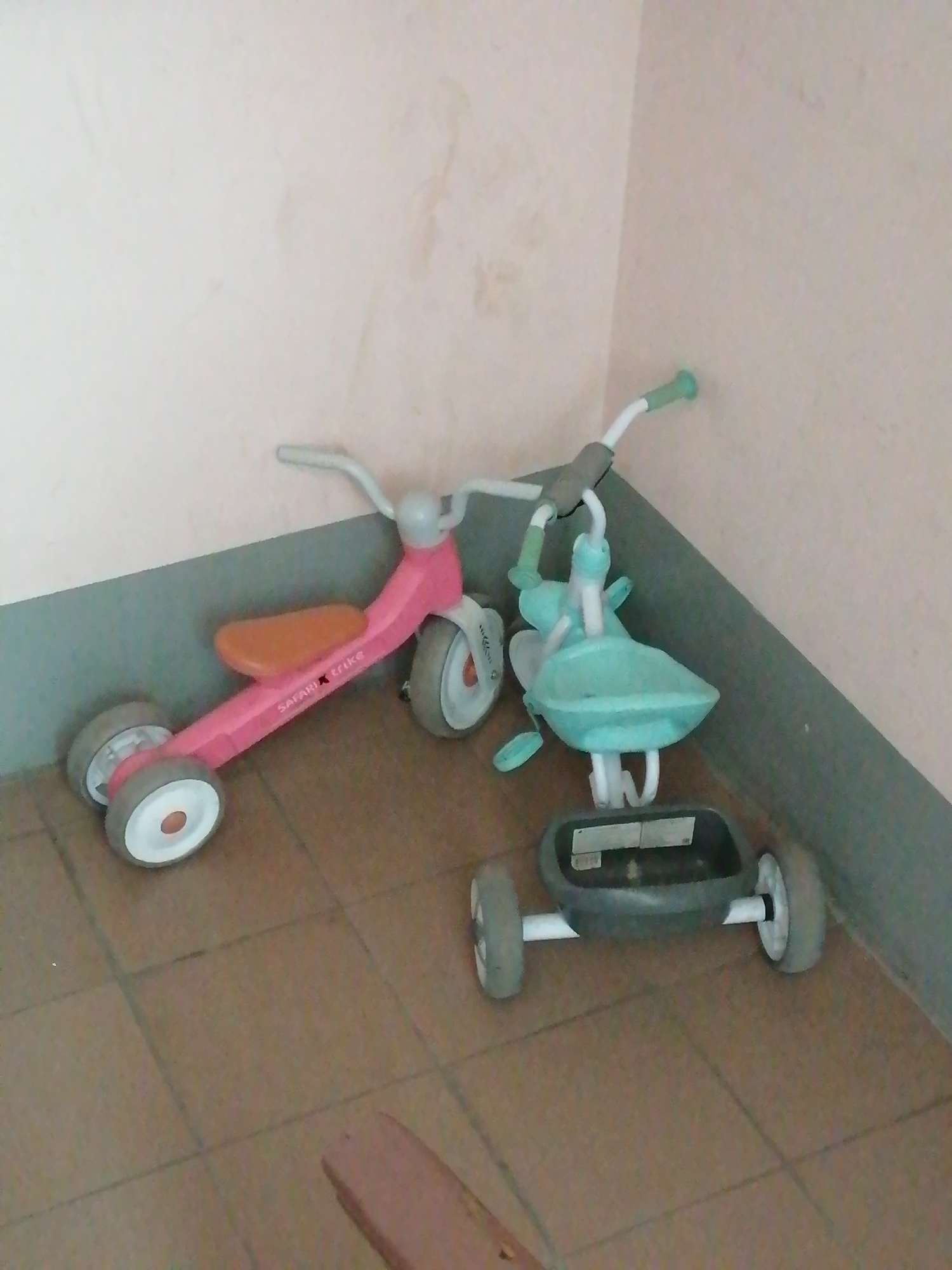 The image displays two children's tricycles positioned in a corner against a wall, likely indoors. The tricycle on the left is primarily pink with an orange seat and white wheels, featuring the text "Safari Trike" on its frame. The tricycle on the right is teal and appears to be a push-along walker or ride-on toy with a tray-like seat and white wheels. The floor is tiled in a terracotta color. The lighting is dim, suggesting an indoor environment. The scene is devoid of people. No specific landmarks or details indicating the city of Rostov-on-Don, Russia, are present in the image.