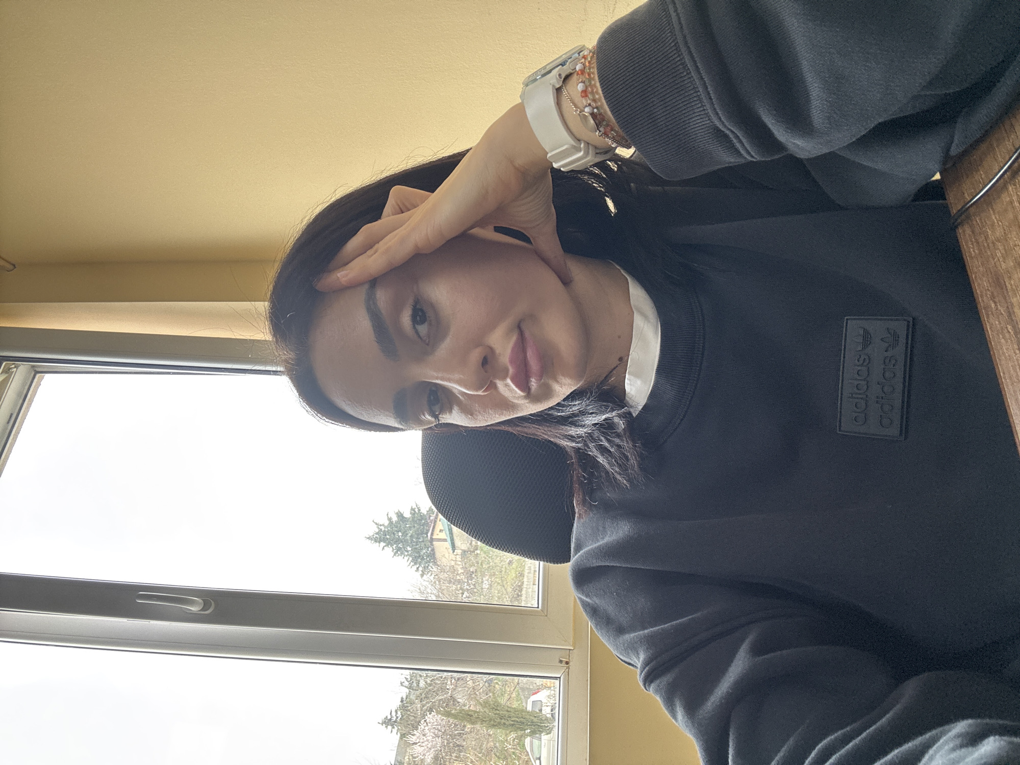 A female individual is depicted in an indoor, chest-up portrait, resting her left hand with fingers on her temple and thumb supporting her jawline. She wears a black crewneck sweatshirt featuring a black Adidas logo patch and a white undershirt. A white digital watch and multi-colored beaded bracelets adorn her left wrist. The background consists of a light yellow wall to the right and a white-framed window to the left. Outside the window, a hilly landscape under an overcast sky features coniferous trees, deciduous trees with light blossoms, a distant residential structure with a red roof, and a white vehicle parked on a lower road. The scene is located in Tbilisi, Georgia.