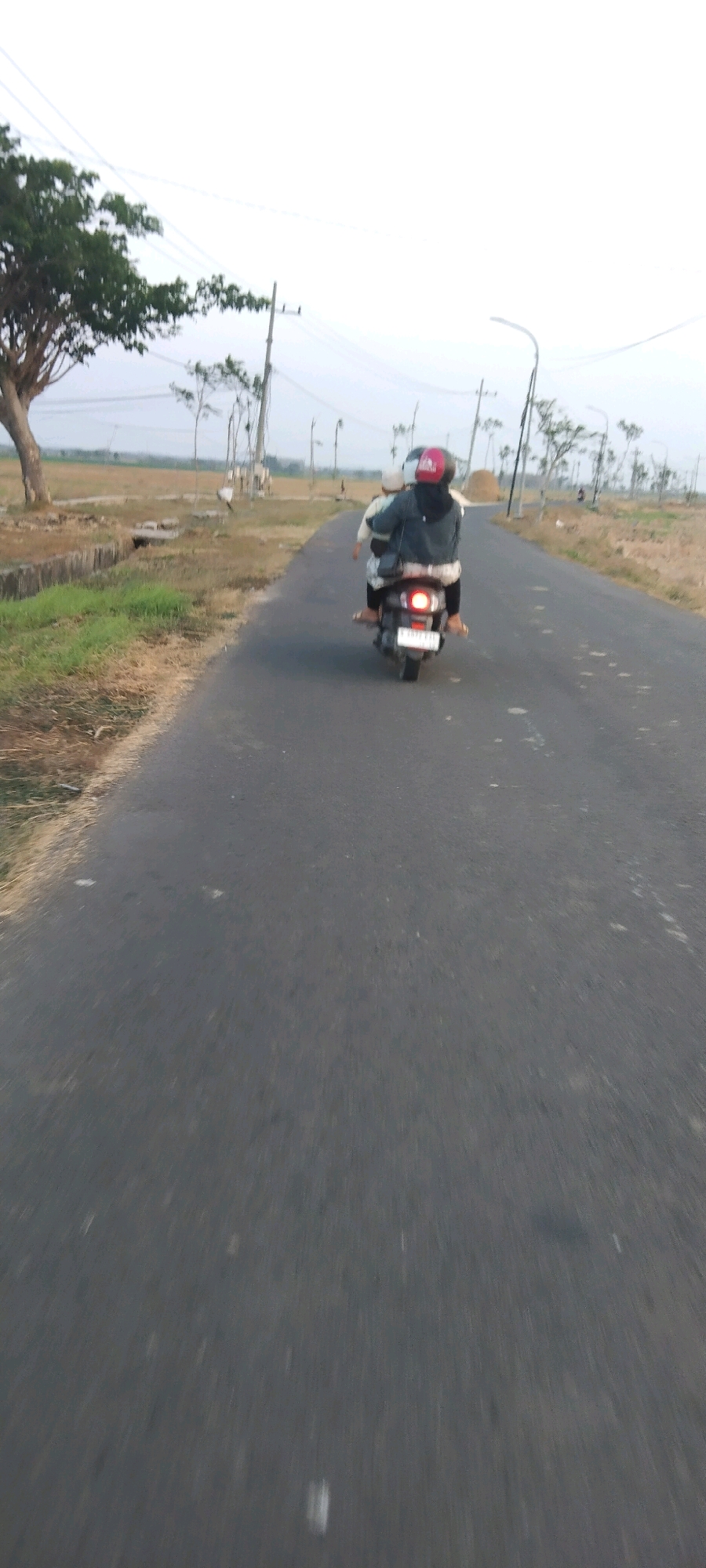 Two individuals are riding a scooter on a paved road in a rural area. The person in front is wearing a dark jacket and a red helmet, while the person behind is wearing a white head covering. The scooter has a white license plate. The road is bordered by dry grass and a concrete drainage ditch on the left. In the background, there are trees, utility poles with wires, and open fields. The sky is overcast. The location is identified as Ngujo, Indonesia.