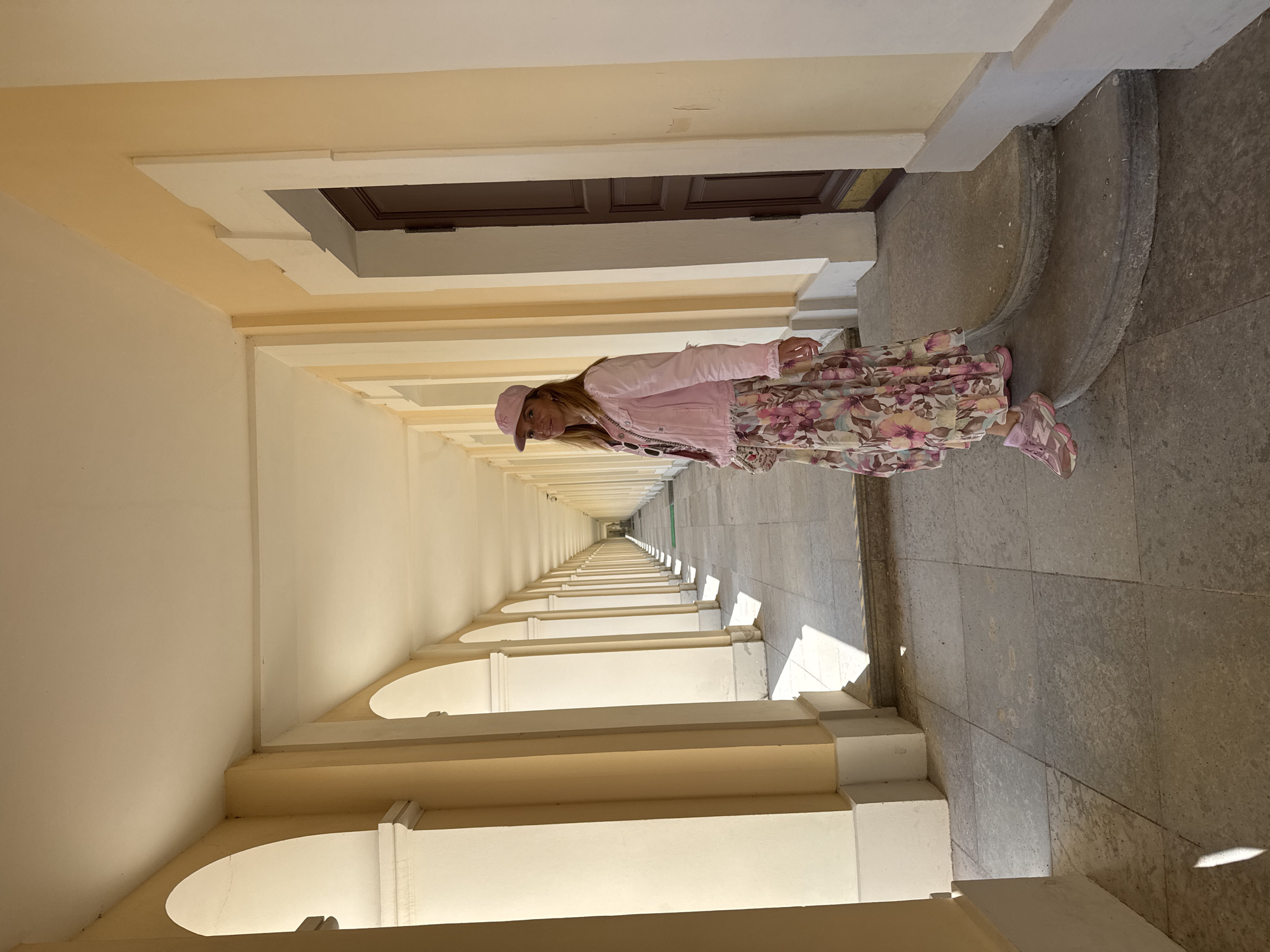 The image shows a woman standing in a long, covered walkway with a repeating architectural pattern of arches and columns. The walkway extends into the distance, creating a strong sense of perspective. The woman is wearing a light pink jacket, a floral patterned skirt with pink and green flowers, and a pink baseball cap. She is also wearing pink athletic shoes. She is looking towards the camera. Sunlight streams into the walkway, casting shadows on the floor. The walls and ceiling of the walkway are painted in light, neutral colors. The scene appears to be taking place outdoors during the daytime, under clear weather conditions. The location is Lomonosov, Russia. There is no visible text in the image. The overall mood of the image is bright and cheerful, with the woman's attire adding a pop of color to the otherwise muted architectural surroundings. The repeating architectural elements suggest a grand or historic location.