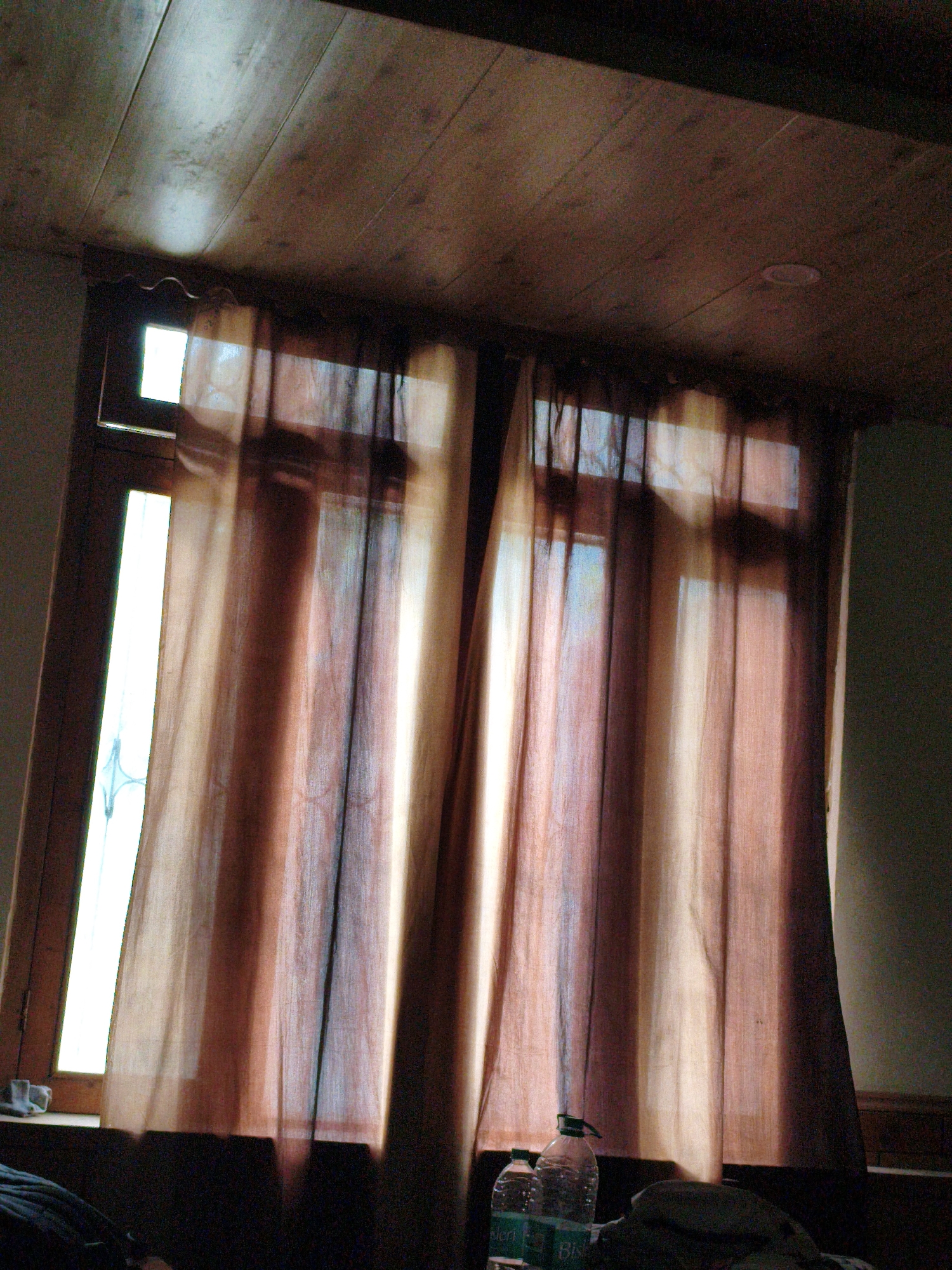 This indoor, eye-level shot captures a room with a wooden ceiling and a window covered by sheer, floor-length curtains. The curtains, in shades of brown and beige, are drawn and allow sunlight to filter through, creating vertical streaks of light and shadow. The window itself appears to be a tall, narrow opening with a wooden frame. The scene is dimly lit, suggesting either early morning or late evening, and the overall mood is subdued and peaceful.
In the foreground, on a surface below the window, are two plastic water bottles. The bottle on the right is larger and has a green cap. The bottle on the left is smaller and has a white cap. Both bottles appear to be standard drinking water bottles. Behind the bottles, a dark, soft object, possibly a piece of clothing or bedding, is visible.
The wooden ceiling is made of planks, some of which are illuminated by a bright light source, likely from the window or an overhead lamp not visible in the frame. The texture of the wood is visible, with grain patterns and some faint marks or blemishes.
The setting is likely a hotel room or guesthouse in Vashisht, India, given the natural light and the presence of water bottles. The time of day is not definitively clear but is suggested to be either dawn or dusk by the quality of light and shadows. There are no people visible in the image. The overall impression is one of quiet domesticity or rest.