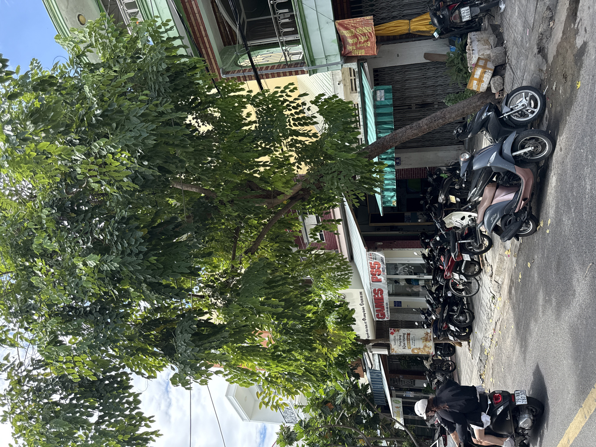 In the city of Nha Trang, Vietnam, a street scene unfolds under a bright, partly cloudy sky. A large tree with dense green foliage dominates the foreground, its branches extending over the sidewalk and a row of commercial establishments. Parked along the curb are numerous motorcycles, a common mode of transportation in the city. On the left side of the image, a person wearing a white helmet and a black jacket rides a black scooter. The scooter's license plate reads "79-NA 287.37".
The buildings lining the street appear to be two or three stories high, with shops at street level. One shop prominently displays signs that read "GAMES" and "PS5". Another sign, partially obscured by the tree, appears to advertise a business called "Yam Kids". The storefronts have awnings, some green and striped, others a solid color. The architecture suggests a blend of modern and older styles, with some buildings featuring balconies and ornate railings.
The pavement is wet in places, indicating recent rain or street cleaning. Scattered yellow leaves lie on the ground near the parked motorcycles. The overall atmosphere is that of a typical urban street in Vietnam, bustling with activity and characterized by the ubiquitous presence of motorcycles.