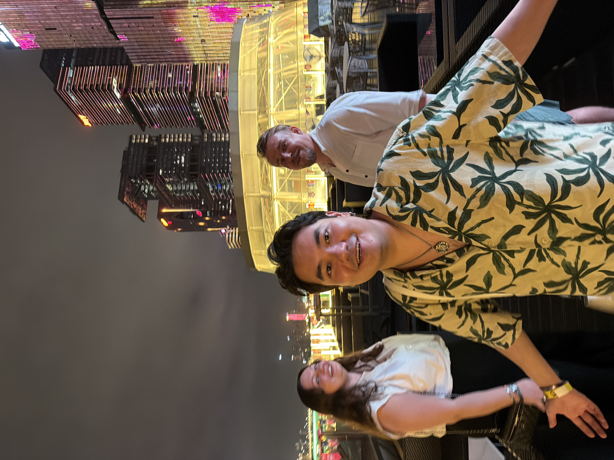 In Haizhu District, China, a nighttime photograph captures three individuals on an elevated outdoor patio, with illuminated skyscrapers forming the backdrop. The person in the foreground, a man with dark hair, is smiling broadly and wearing a light-colored, short-sleeved shirt adorned with a green leaf pattern. His right hand is extended towards the camera, and he is wearing a necklace with a circular pendant and a yellow wristband. To his left, a woman with long, wavy brown hair, wearing a white sleeveless top and black pants, is laughing. She has a silver bracelet on her left wrist and a black purse slung over her shoulder. Behind the man in the foreground and to the right, a man with short blond hair and a stubbled beard is wearing a white button-down shirt. He is looking slightly to his left. The patio features tables and chairs. In the background, the city skyline is dominated by tall, modern buildings, many of which are brightly lit with pink and yellow lights. A circular, illuminated structure is visible in the mid-ground.