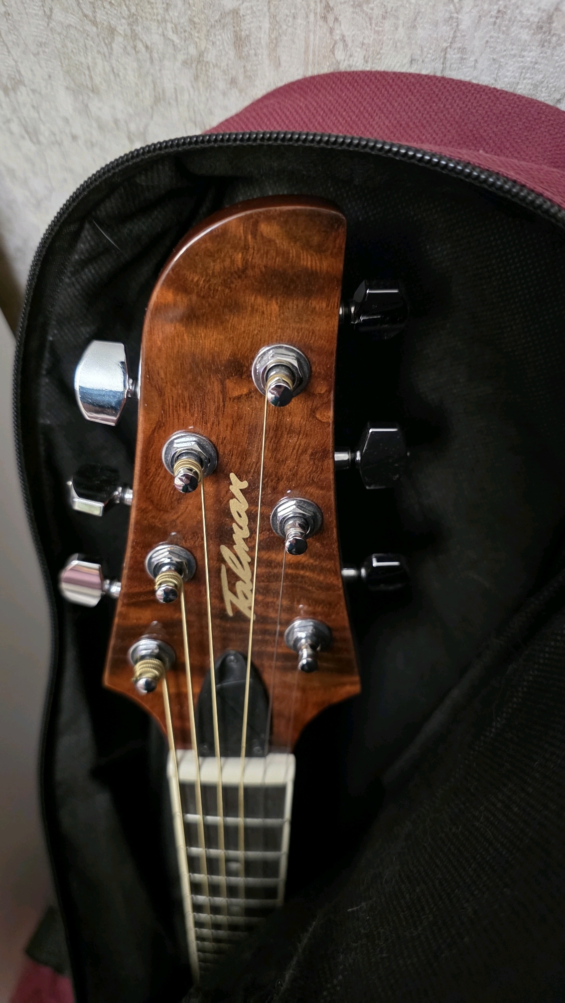 A close-up, eye-level shot shows the headstock of an acoustic guitar resting inside a dark-colored guitar case, with the burgundy outer fabric of the case visible at the top. The guitar's headstock is made of polished, reddish-brown wood with a flame pattern, featuring six tuning pegs. The tuning pegs on the left are silver, while those on the right are black. A stylized script logo that reads "Talman" is inlaid on the wood of the headstock. Four light-colored strings run down from the tuning pegs towards the fretboard, which is partially visible and appears to be made of wood with white fret markers. The background consists of a textured, light-colored wall. There are no people or animals present.
