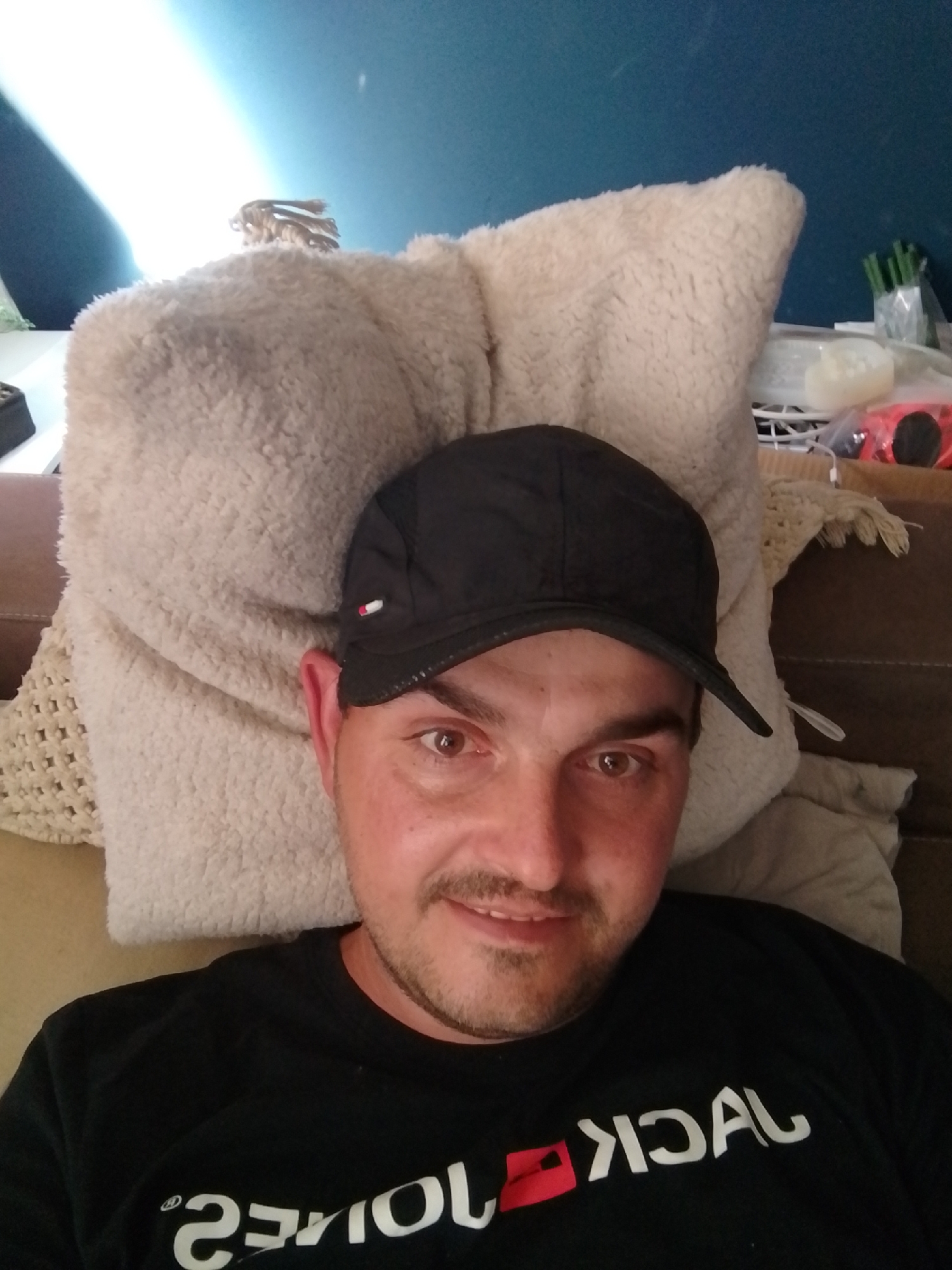 A man wearing a black baseball cap and a black t-shirt with white text lies on a sofa, propped up by a large, fluffy, cream-colored pillow. The man is looking directly at the camera, with a slight smile. He has light brown eyes and stubble on his chin and upper lip. The t-shirt has text that appears to be reversed, with the word "JACK" clearly visible. In the background, a blue wall is visible, with a diagonal beam of light shining from the left side of the frame. Some items are visible on a shelf behind the man, including a vase with green stalks and a red object that appears to be a toy car. The scene appears to be indoors, possibly in a living room. The lighting suggests it is daytime.