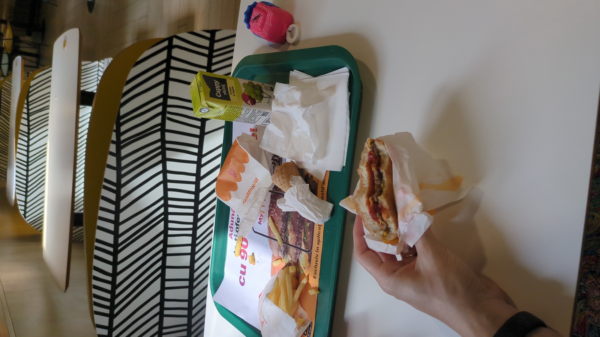 A close-up, eye-level shot shows a person's hand holding a partially eaten McDonald's cheeseburger over a white table. The burger is wrapped in white paper, with some grease stains visible. In the background, a green McDonald's tray contains a small box of Cappy apple juice, a bag of French fries, an empty McNuggets bag, a crumpled napkin, and a small toy with a spider web design. A large McDonald's placemat with an image of a burger is also visible. The setting appears to be a McDonald's restaurant in Chișinău, Moldova, indicated by the placemat's text and the presence of McDonald's branding. The background features stylized seating with black and white patterns.