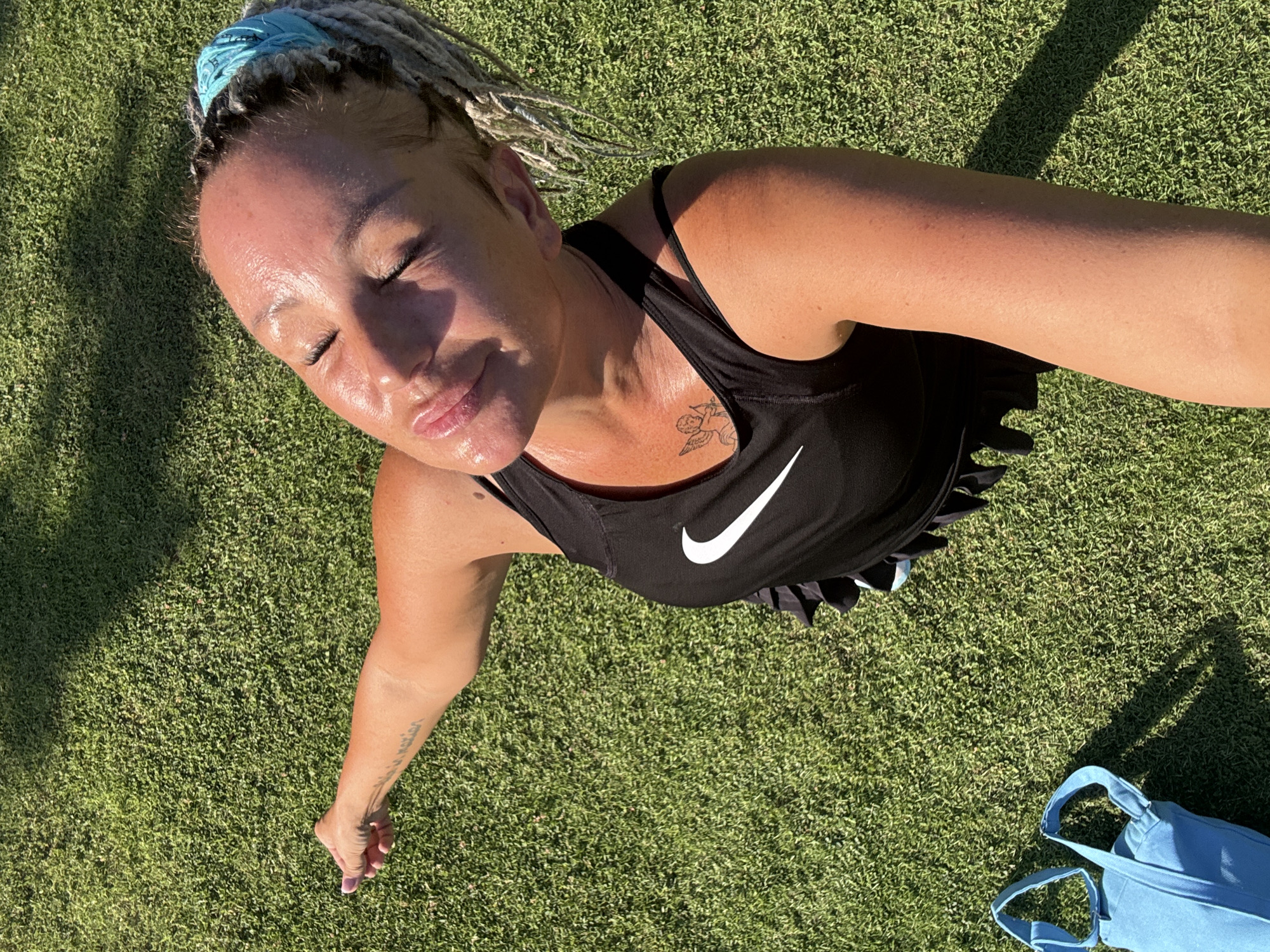 A high-angle outdoor selfie captures a woman on a sunny day. She has light-colored dreadlocks tied back with a light blue bandana. Her face is sweaty and flushed, with her eyes closed and a soft smile, conveying a sense of relaxation or enjoyment of the sun. She wears a black sports tank top featuring a prominent white Nike swoosh logo on the chest. A small, winged tattoo is visible on her upper right chest, and a script tattoo adorns her left forearm.
The background is entirely composed of vibrant green grass, suggesting an open field, park, or garden. Strong, direct sunlight casts distinct shadows, indicating a bright, clear day, likely in the morning or late afternoon. A light blue fabric bag is partially visible on the grass in the lower right corner of the frame. The overall impression is one of physical activity or relaxation in a warm, sunny environment, consistent with Kuta Selatan, Indonesia.