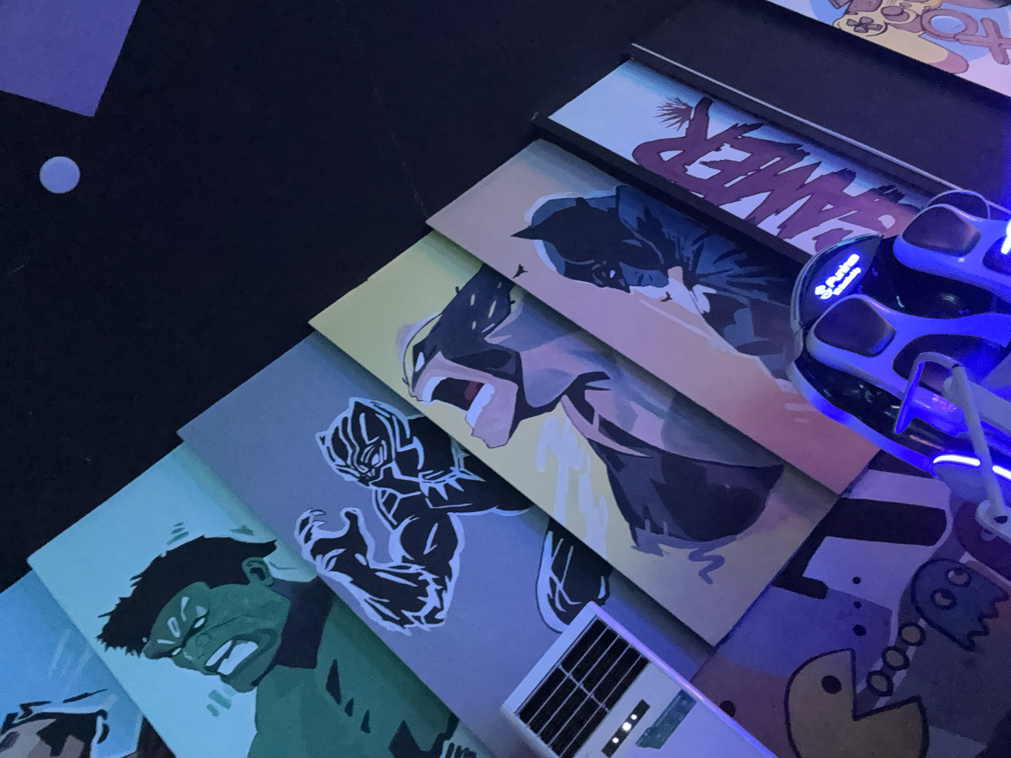 This media file, captured in Abuja, Nigeria, displays a collection of pop culture art panels and gaming equipment arranged diagonally on a dark surface, likely a table or desk. The scene is dimly lit, with a prominent blue glow emanating from a gaming headset on the right, suggesting an evening or night setting.
Several rectangular art pieces are visible, featuring stylized comic book and video game characters. From left to right: one panel depicts a green-skinned Hulk with an angry expression. Next, Black Panther is shown in his iconic suit against a grey background. Following this, another dynamic illustration of Black Panther with an open mouth is visible on a yellow/beige background. Adjacent to this is a panel featuring a silhouette of Batman. Further right, a white panel displays the stylized word "GAMER" in purple text. The bottom right reveals parts of another panel with classic video game characters, including Pac-Man and ghosts.
A black and blue gaming headset, illuminated with a bright blue light and bearing the text "Pulse," sits prominently on the right side, partially covering the "GAMER" panel. Below the Black Panther and Hulk panels, a small white electronic device with a black grid and three illuminated dots is visible. A faint, light blue circular reflection or object is seen in the top-left corner of the dark surface. The overall impression is a vibrant, modern gaming or entertainment setup, emphasizing popular culture themes.