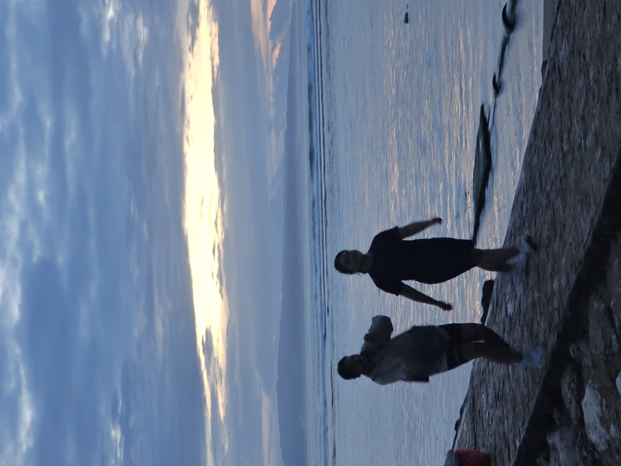 Two people stand near the water along a concrete structure. The sky is filled with clouds and a bright glow, possibly from the setting sun. The image was captured in Denpasar, Indonesia.