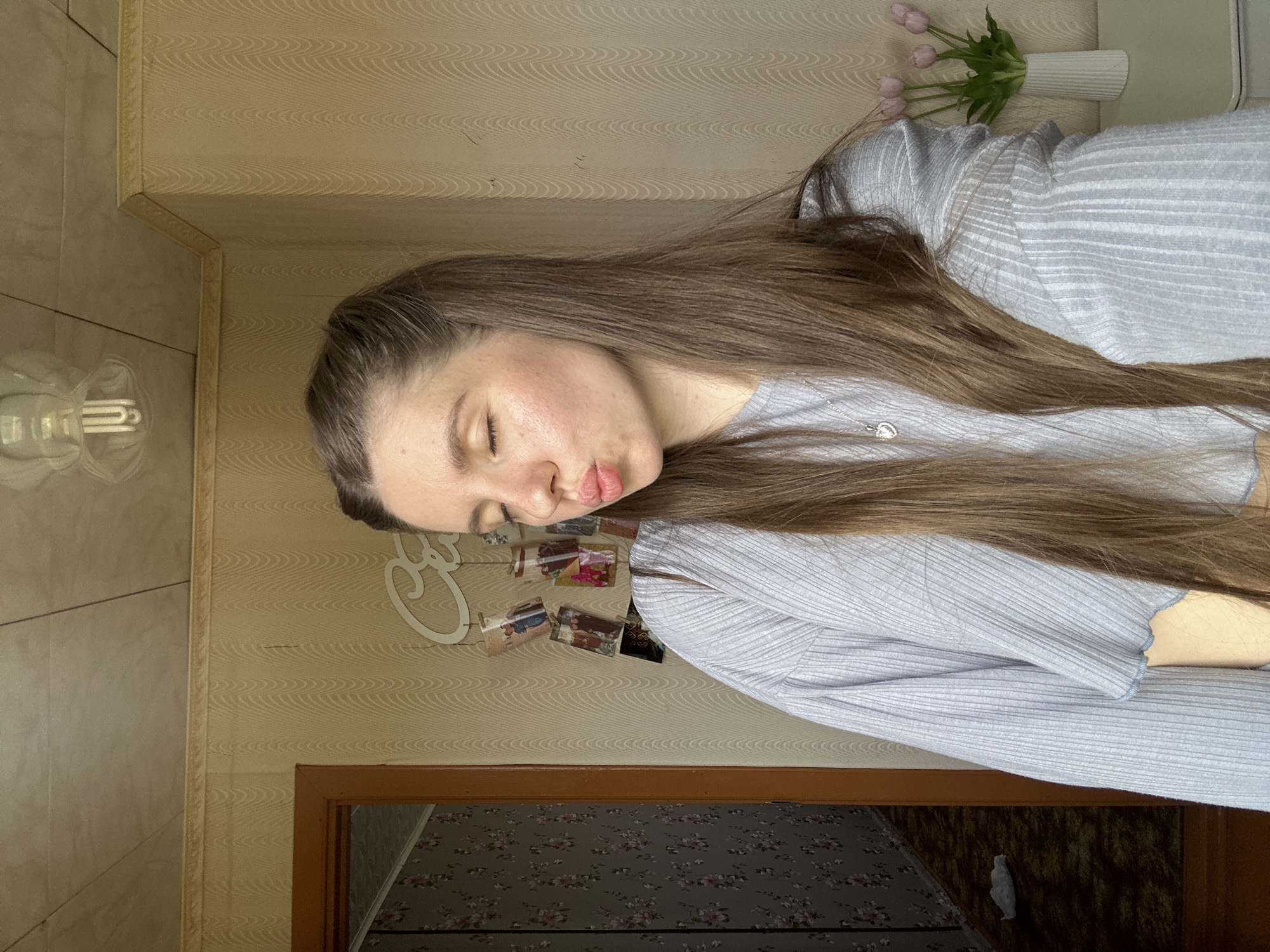 A young woman is photographed indoors. She has long brown hair and is wearing a light blue ribbed shirt. Her eyes are closed, and she has pursed lips. Behind her are light beige patterned walls. On the wall, there is a decorative word and several small pictures. To her right is a vase with pink tulips. Below her is a partially visible doorway with a floral-patterned floor visible within. The location is unknown; there is no information that the scene is in the city of Мещёры, Russia.