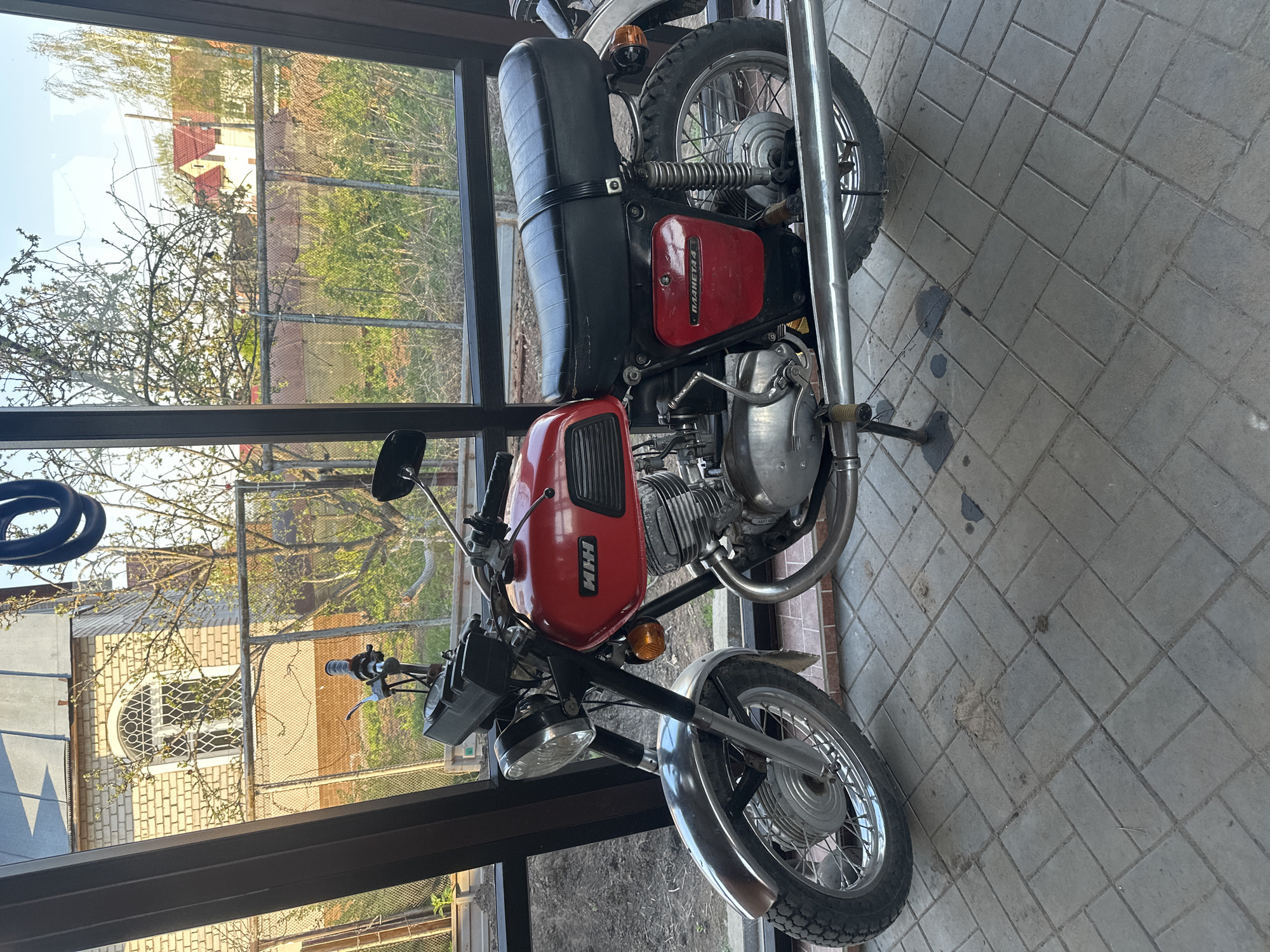 The image shows a red and black motorcycle parked indoors. The motorcycle has the Cyrillic letters "ИЖ" on the gas tank. The motorcycle is parked on a paved surface within a structure with dark-framed windows. Through the windows, a view of a residential area is visible, including buildings, trees, and fences. There is no evidence in the image to confirm that it is located in the city of Chaikovsky, Russia.