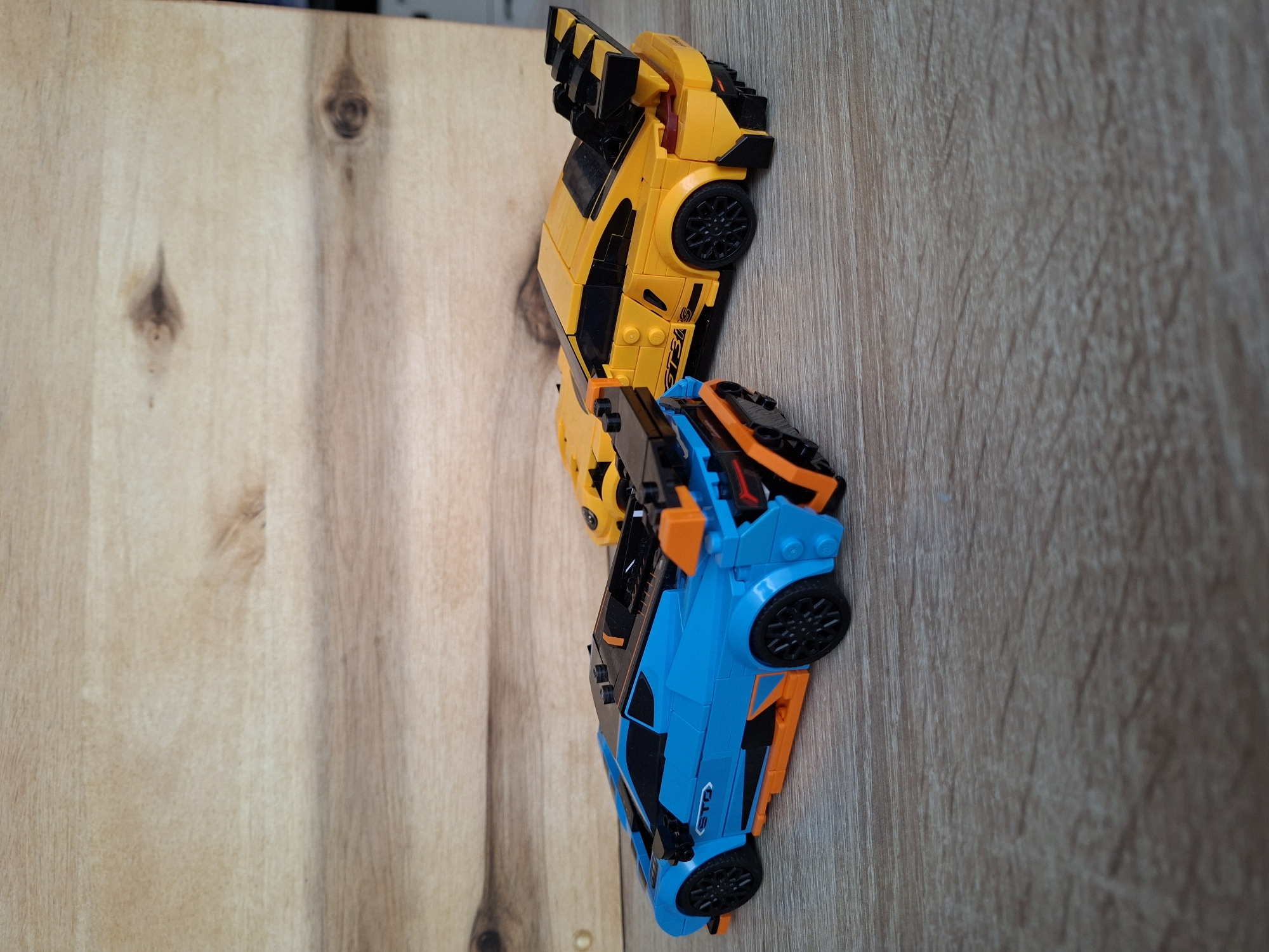 The image depicts two Lego toy cars on a wooden surface. The car in the foreground is blue and orange with black wheels and detailing. The car behind it is yellow with black accents, including a large rear spoiler and black wheels. The background is a light-colored wooden plank with visible grain and knots. There is no indication of the city of Baden-Baden, Germany, or any people present. The scene is a still life composition of toy vehicles.