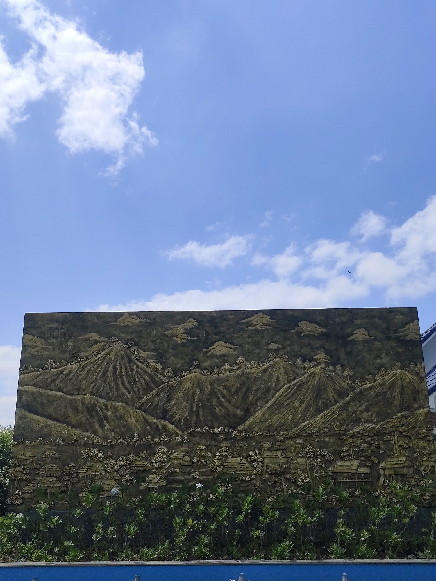 The image displays an outdoor scene dominated by a large, dark-toned relief mural under a bright blue sky with scattered white cumulus clouds. The mural, which occupies a significant portion of the lower frame, depicts a detailed mountainous landscape. It features several prominent, textured peaks, stylized clouds floating above them, and numerous traditional-style houses nestled amidst trees at the base of the mountains, suggesting a village setting. The artistic style of the mural evokes a sense of local culture, consistent with the location context of Lạc Dương, Vietnam, likely representing a highland or rural community.
In front of the mural, a row of vibrant green bushes and smaller plants forms the immediate foreground, with a solid blue structure visible along the very bottom edge of the frame. Above the mural, the clear sky is dotted with a few fluffy white clouds. A small, dark bird can be seen in flight in the upper right portion of the sky. On the far right, a sliver of a building with a blue roof is visible, peeking above the mural. The scene is bathed in natural daylight, indicating a sunny, clear day. No human activity or visible text is present in the image.
