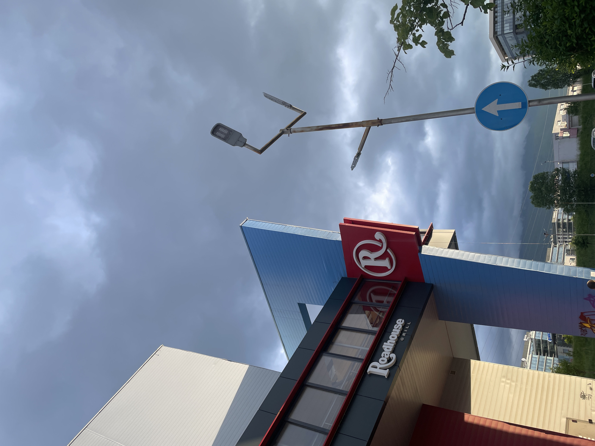 The image is taken at an upward angle, capturing architectural elements against a cloudy sky. Dominating the foreground is a "Roadhouse Grill" restaurant, identified by its signage featuring the restaurant's name and logo ("R" in a circle). The building incorporates geometric forms in blue, red, black, and white. A streetlight with a modern LED fixture and a rusted support structure is visible. Additionally, a blue circular sign with a white arrow pointing left is present. In the background, other buildings and some greenery are discernible. The location is Sofia, Bulgaria.