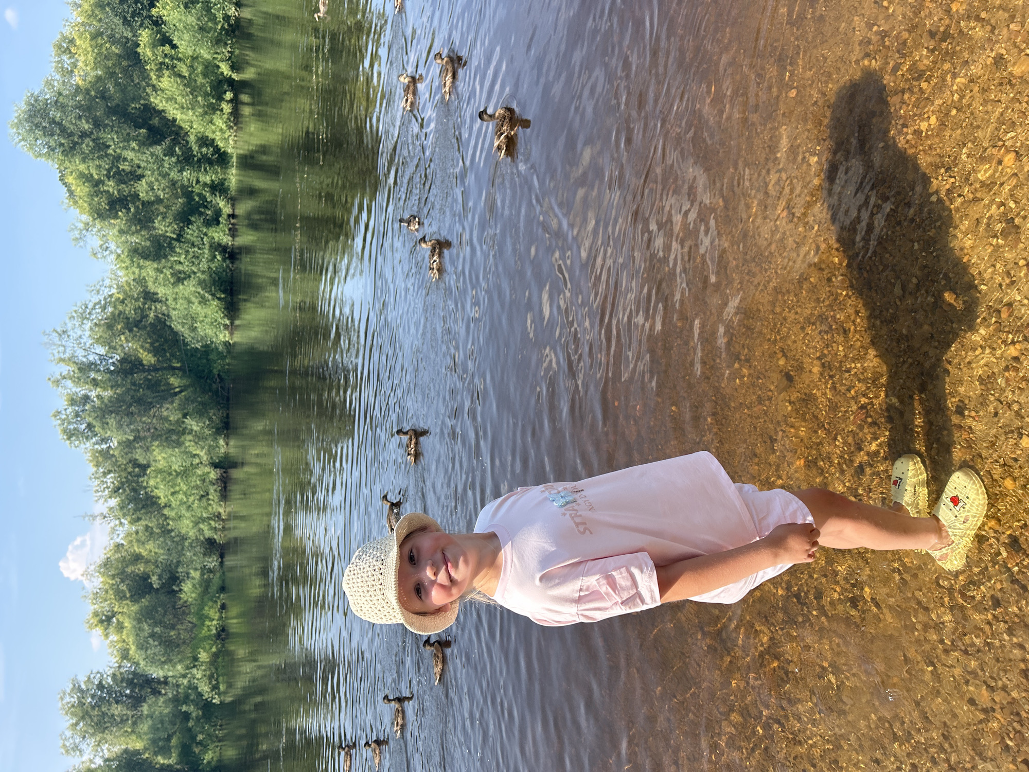 The image depicts a young girl standing at the edge of a body of water, likely a river, in Krasnovishersk, Russia. She is wearing a light-colored bucket hat, a pink t-shirt with a graphic, and yellow clogs. Several ducks are swimming in the water behind her. The background consists of a dense line of green trees and a blue sky with some clouds. The water is calm with ripples, and the bottom is visible and rocky where the girl is standing. The lighting suggests it is daytime, likely midday given the shadows.