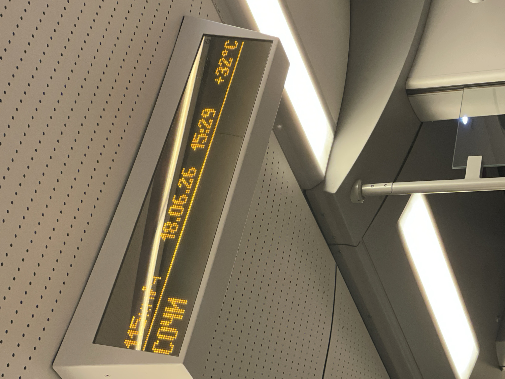 This image captures a digital display inside what appears to be a train or a similar public transportation setting in Monastyr, Russia. The display is oriented vertically and shows information in bright yellow LED characters against a dark background.
The visible text on the display includes a date and time, "18.06.25 15:29," and a location, "СОЧИ" (Sochi). It also shows a temperature reading, "+32°C". The setting is dimly lit, with bright overhead lights creating strong reflections on the display and surrounding surfaces. The wall behind the display is textured with a pattern of small, regular dots. The overall impression is of a functional, modern interior, likely within a travel context. There are no people visible in the image. The time of day is mid-afternoon, and the temperature indicates warm weather.