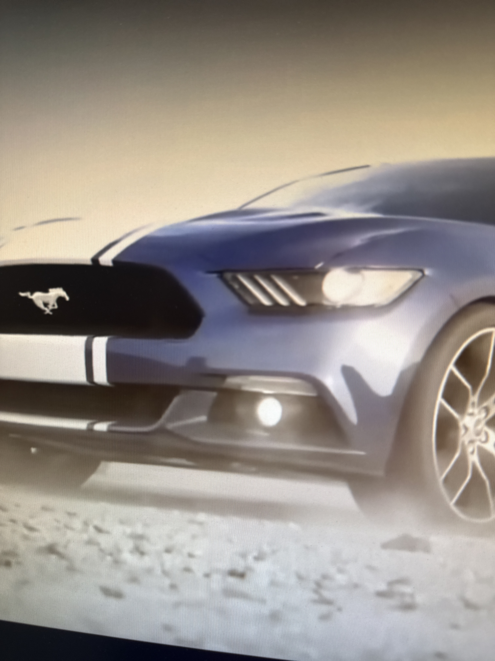 The image displays a close-up, low-angle view of a blue Ford Mustang sports car. The car is positioned on a dusty, unpaved surface, possibly a dirt road or an open field. The background is a hazy, pale sky. The car's front is prominent, showing its grille with the iconic Mustang horse emblem, distinctive headlights, and racing stripes on the hood. The front bumper and a portion of the driver's side wheel are also visible. The overall lighting suggests an outdoor environment, likely during the daytime. There are no people or any other discernible objects or landmarks that would indicate the specific location within Netanya, Israel. The scene is static, with no action or interaction depicted.