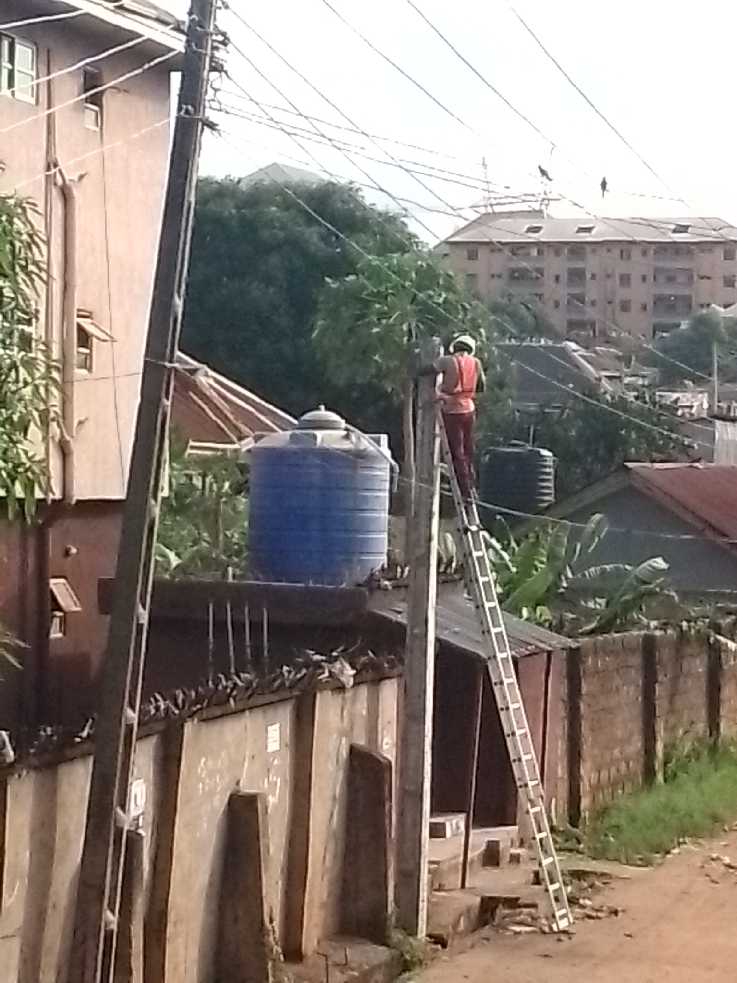The image captures an outdoor scene, likely a residential or semi-urban area, dominated by a utility pole and a person working on it. In the foreground, a tall wooden utility pole stands prominently on the left, with numerous electrical or communication wires stretching across the frame against a bright, overcast sky.
A person, seen from behind, is positioned high on a long, silver extension ladder that leans against this utility pole. The individual wears a light-colored hard hat and a bright orange reflective vest over dark clothing, suggesting they are a worker engaged in maintenance or installation. Their body faces away from the viewer, concentrating on the pole or wires. The bottom of the ladder rests on uneven ground or a raised concrete surface near a small structure.
In the mid-ground, behind the utility pole, several structures and objects are visible. To the left, part of a multi-story building with small rectangular windows is seen. Behind the worker and the utility pole, two large water tanks are present: one prominent blue, cylindrical tank and another, darker, ribbed tank partially obscured. Further back, more residential buildings with tiled or corrugated roofs are scattered amidst dense green foliage and trees. A long, textured concrete wall, possibly a retaining wall or fence, runs horizontally across the lower middle section of the image, with what appear to be birds perched along its top edge.
In the far background, a large, multi-story building with numerous windows and a flat or low-pitched roof is discernible under the network of wires, hinting at a more densely populated area. The lighting suggests it is daytime, possibly late morning or early afternoon, with diffused sunlight. No discernible text is clearly visible on any of the objects or surfaces within the image. The scene conveys a sense of everyday work taking place amidst typical urban infrastructure in what appears to be a warm climate.