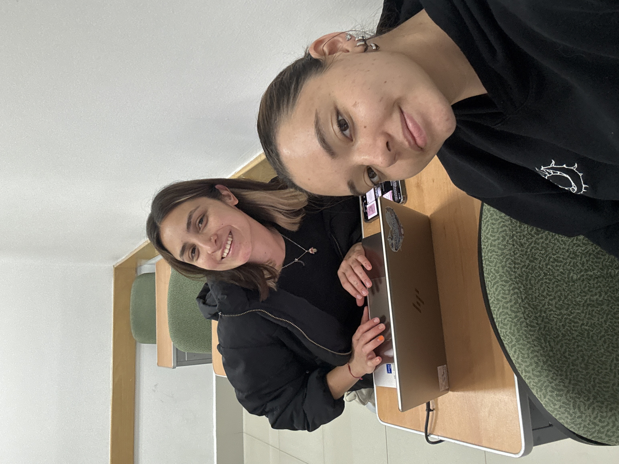 The scene takes place in an indoor classroom or study room in San Andrés Cholula, Mexico. Two young women are present. In the foreground, positioned to the right, a woman with dark hair, wearing a black hoodie with a white embroidered design, looks directly at the camera with a slight smile. She has multiple earrings, including hoops and studs.
To her left and slightly behind, another woman with brown hair and lighter streaks smiles towards the camera. She is dressed in a black puffer jacket over a black top and wears a silver necklace with a small, light-colored charm. Her hands are positioned above an open, metallic-colored HP laptop, which displays "Intel Core" and "Corazon" stickers. A mobile phone is visible on the desk beside the laptop. Her left hand shows one orange-painted fingernail and a red string bracelet.
The environment consists of light wooden desks with white edges and green, patterned upholstered chairs. Two empty chairs are visible in the background. The walls are plain white, with a light wooden baseboard along the lower section of the back wall. The room is brightly and evenly lit.