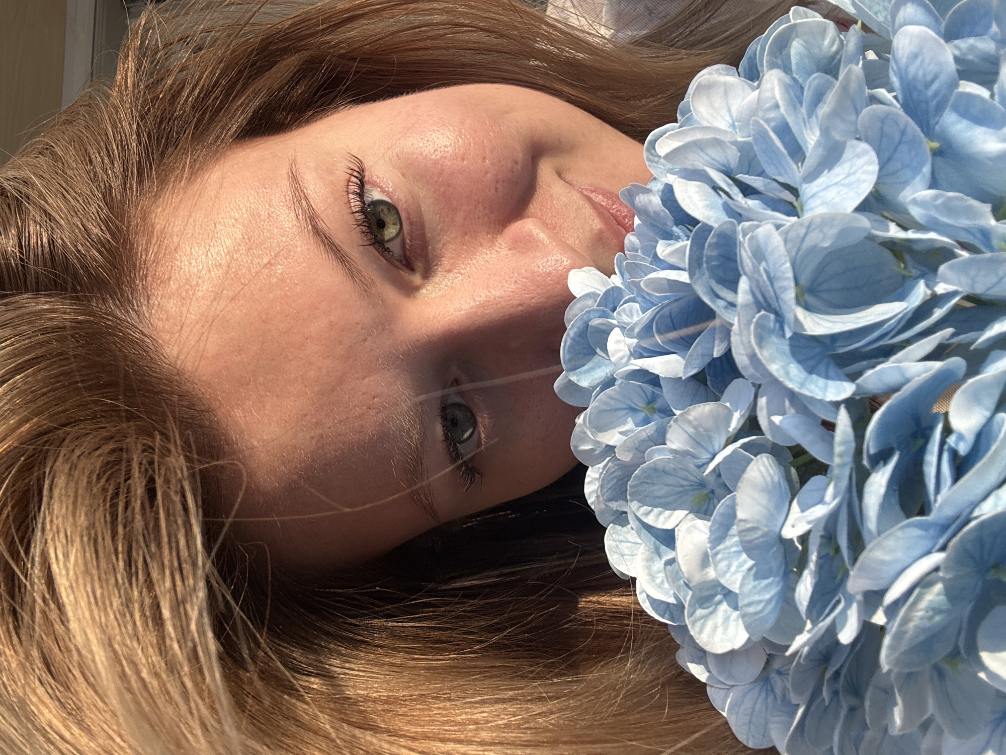 A close-up, slightly rotated portrait shows a fair-skinned woman with light brown hair next to a bouquet of blue hydrangeas. The woman's face is partially shaded by the bright sunlight. Her eyes are a mix of green and brown, and her hair is predominantly blonde with darker roots. The bouquet of blue hydrangeas sits to the right of her face, covering part of her cheek and chin. The background is indistinct, but appears to be outdoors, with indirect sunlight. There's no information to confirm the location is in Chaikovsky, Russia.