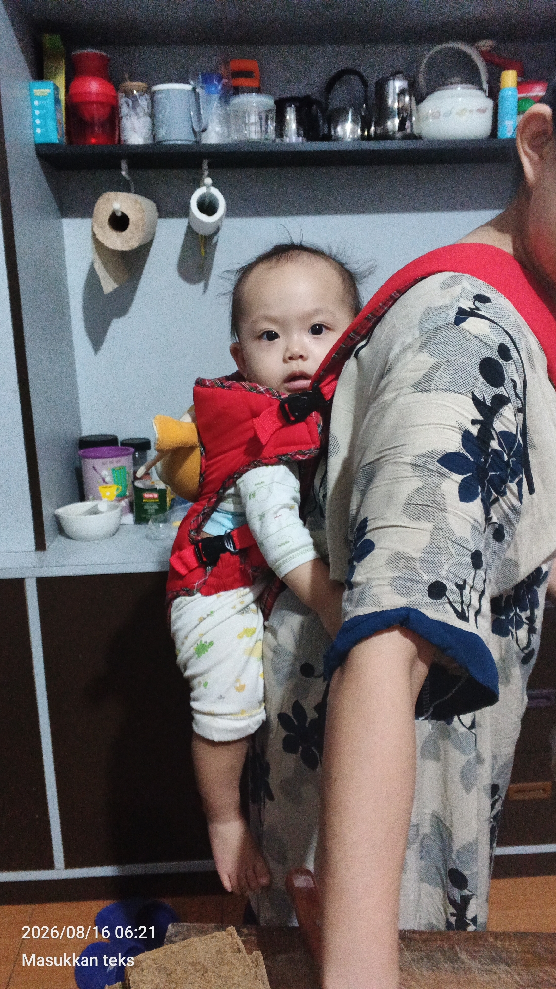 A baby is being carried on the back of an adult in a red carrier. The baby is wearing a white outfit with colorful patterns and a red bib. The adult is wearing a patterned shirt. The background shows a shelf with various items, including containers and a kettle, and a wall with two rolls of toilet paper hanging from hooks. A timestamp and text are visible in the bottom left corner of the image. The scene appears to be indoors, possibly in a home or kitchen. The image was captured on August 16, 2026, at 06:21.