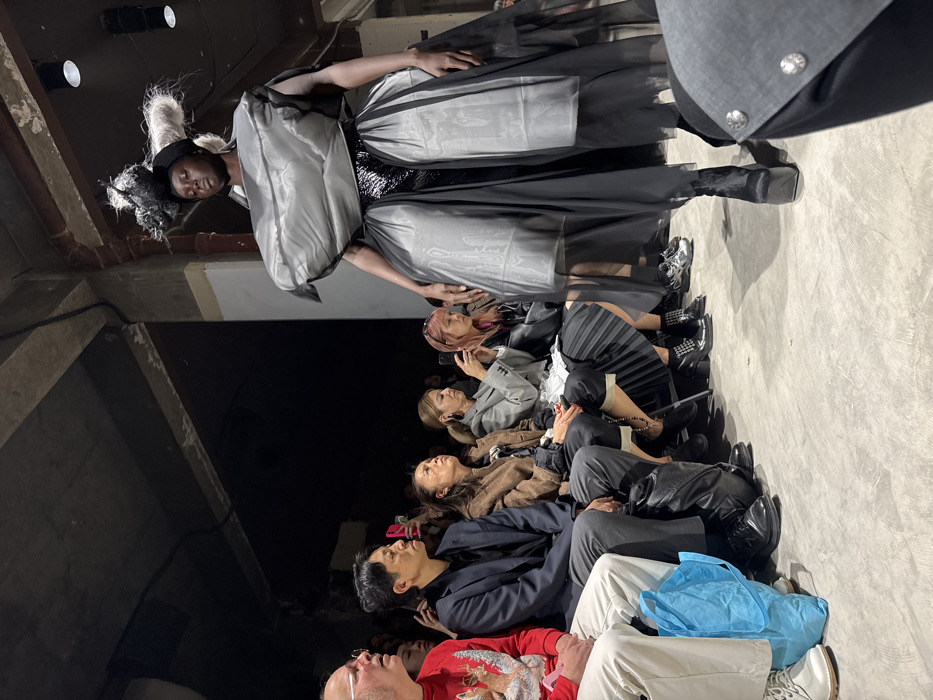 The image captures a fashion presentation in an industrial-style venue located in Paris, France. A dark-skinned model, positioned right of center, walks on a grey concrete floor. The model wears an elaborate ensemble consisting of a multi-layered headpiece with black and white textured elements, a voluminous, structured grey padded top covering the torso, a shiny black mid-section, and a long, flowing black skirt with two prominent, grey, rectangular padded panels. Black platform boots complete the look. To the left, a front row of spectators is seated, observing the model. From left to right, individuals include a male wearing glasses and a red sweater with a reindeer motif, a male in a dark suit jacket and black shirt, a female in a brown jacket and dark pants, a female in a grey blazer over a pleated dark skirt photographing with a smartphone, and a female with pink hair also holding a smartphone. A light blue reusable bag rests on the floor in front of the leftmost spectator. The background is dark, revealing concrete architectural elements, exposed pipes, and recessed spotlights.