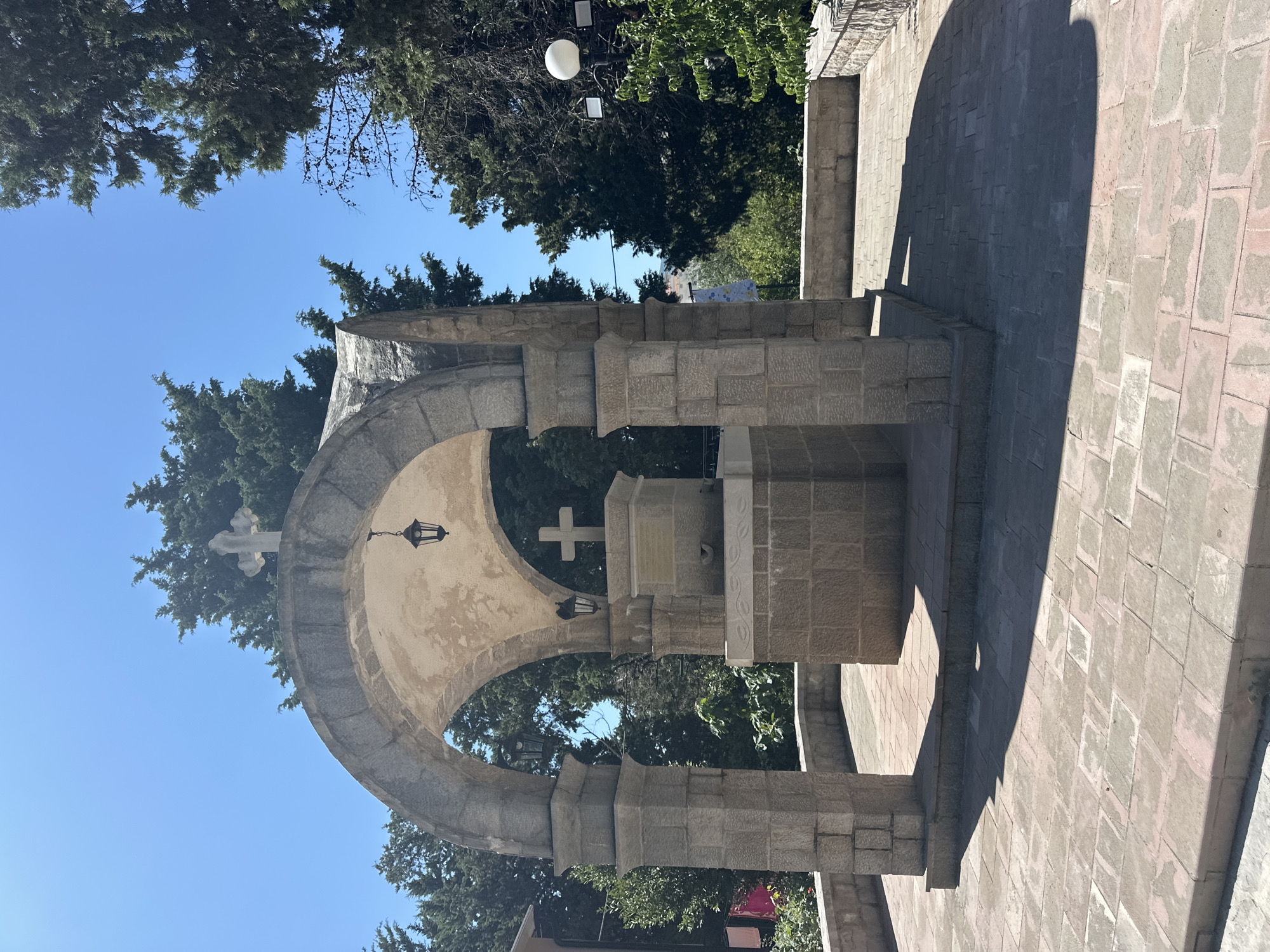 The image shows a stone structure with an arched roof supported by columns, situated outdoors in what appears to be a public space. A cross is visible on top of the structure. There are trees and a clear blue sky in the background. The ground is paved with stone tiles, and there are shadows cast by the structure. The overall impression is that of a monument or religious site. The location is identified as Boreti, Montenegro.
