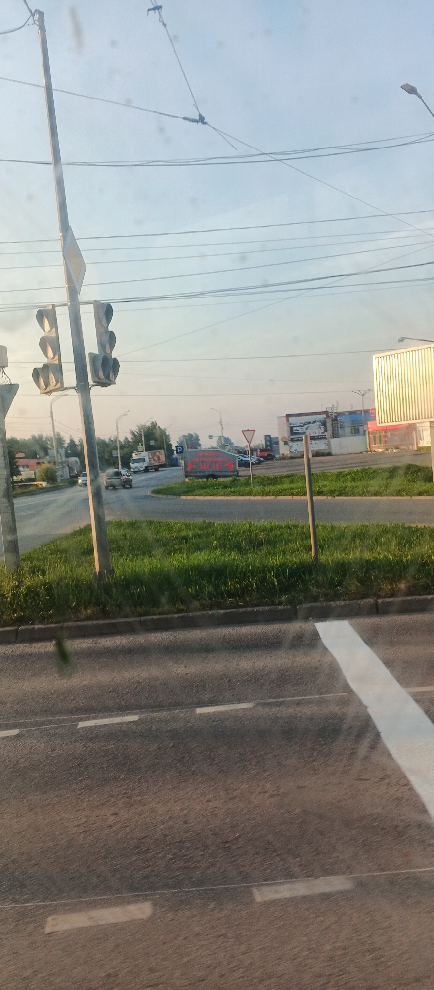The image depicts a view from a vehicle at a traffic light intersection in Izhevsk, Russia. A traffic light with three lamps, two of which are illuminated green, is prominently visible on the left. A signpost with a yellow diamond shape is attached to the traffic light pole. In the background, a roundabout is visible, with a truck and several cars navigating through it. A billboard with red lettering is also present, though the text is mostly illegible. The scene is characterized by a bright, overcast sky and a lush green grassy area separating the road from the background. The road surface shows white lane markings and a pedestrian crossing. The overall impression is of a typical urban street scene during daylight hours.