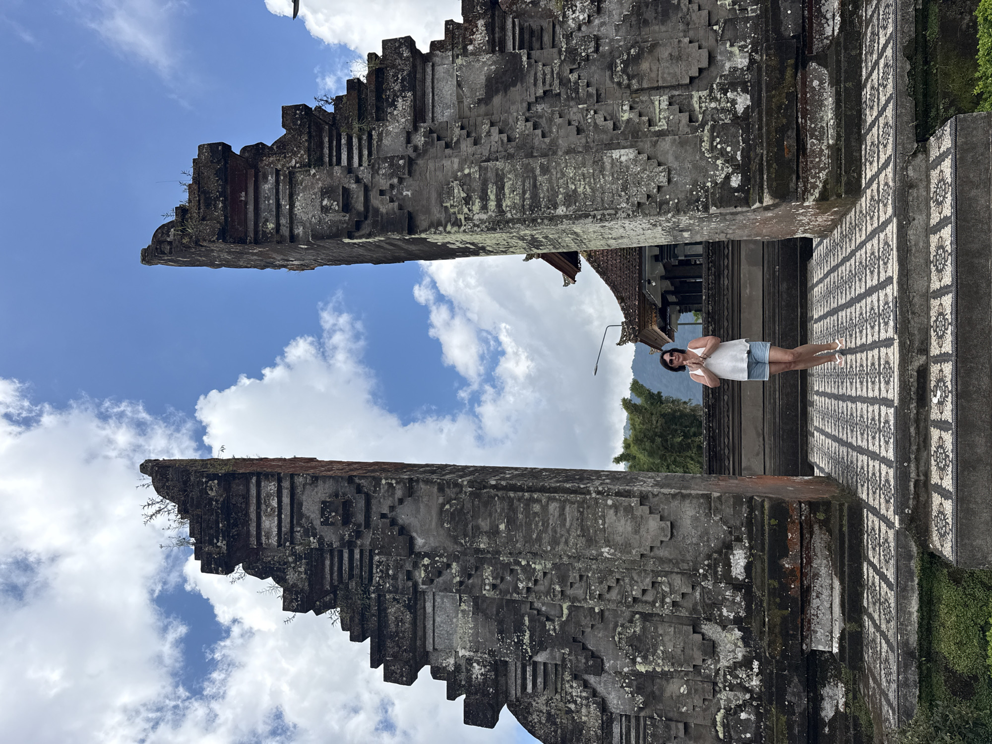 The image depicts a vertical view of a person standing at the Handara Gate in Baturiti, Bali, Indonesia. The gate is composed of two tall, gray stone structures with a patterned, tiered design. The sky visible through the gate is blue with scattered white clouds. A woman, wearing a white top and denim shorts, stands in the gate. Vegetation is visible in the background and to the right. The walkway beneath the gate is tiled with a black and white patterned surface.
