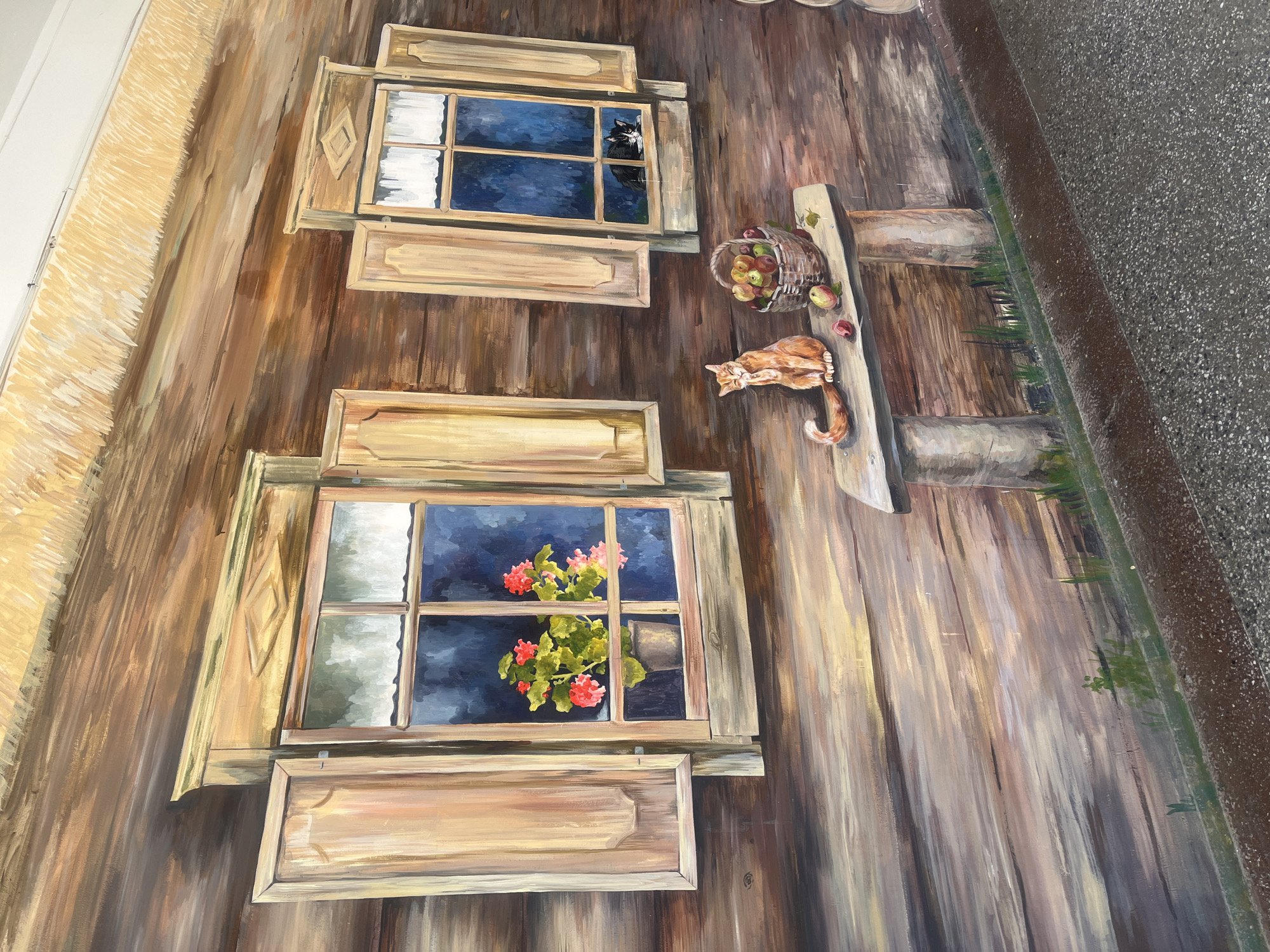 This is a detailed photograph of a mural painted on the exterior wall of a building, likely in the city of Hrodna, Belarus. The mural depicts a rustic scene set against a wooden log structure. Two windows with open shutters are visible, one larger than the other. Inside the larger window, a pot of vibrant red and pink flowers is displayed. The smaller window shows two cats; one is black and white, peeking out, and another, a black cat, is nestled inside. Below the windows, a wooden bench rests on two thick supports. An orange tabby cat sits on the bench, grooming itself. Beside the cat is a woven basket overflowing with apples, with a few loose apples scattered around the bench. The mural is characterized by its realistic, painterly style, with detailed rendering of wood textures, light, and shadow. The ground in front of the mural is a textured, dark gray surface, possibly asphalt or concrete. The mural appears to be located outdoors, possibly along a pedestrian walkway or public space in Hrodna.