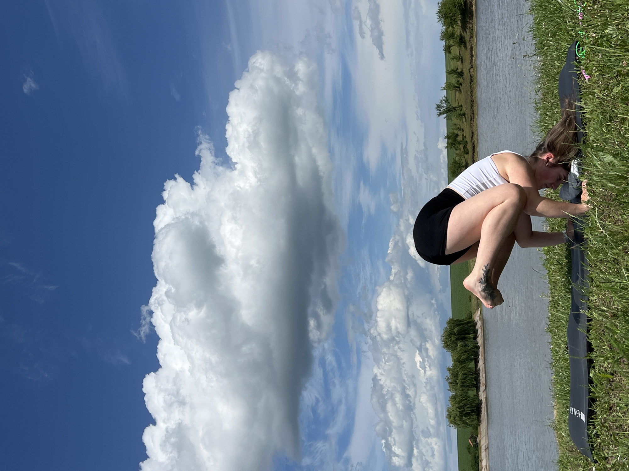 A woman is performing a yoga pose, specifically a crow pose, on a yoga mat outdoors. She is wearing a white tank top and black shorts. Her face is obscured as she is in a bent position with her head close to the ground. Her legs are bent, and her feet are tucked close to her body. Her arms are supporting her body weight. The yoga mat is black and placed on a grassy area. In the background, there is a body of water, likely a river or lake, with trees lining the opposite bank. Beyond the trees, there are green fields. The sky is blue with scattered white and grey clouds. The image appears to be taken on a sunny day. The location is identified as Borychivka, Ukraine.