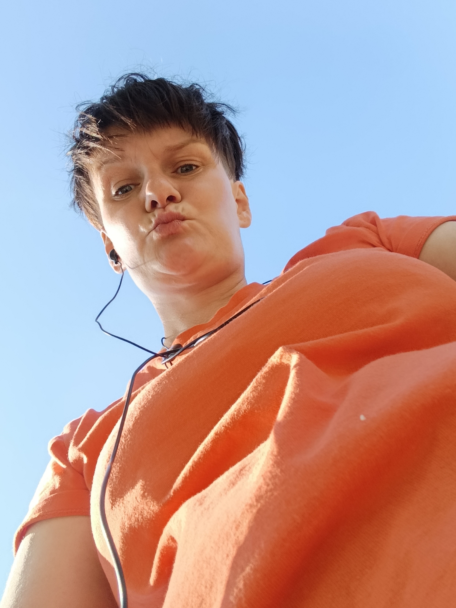 The media file shows a low-angle, upward shot of a person outdoors against a clear blue sky. The subject has short, dark hair and is wearing an orange t-shirt and black earbuds. Their face is turned towards the camera with a playful, puckered expression. The bright sunlight suggests it is daytime, likely a sunny day. The perspective and lighting emphasize the subject's face and the texture of their shirt. There are no visible text elements in the image. The setting appears to be an open outdoor space, given the vast expanse of sky. The location context provided is Фоки, Russia.