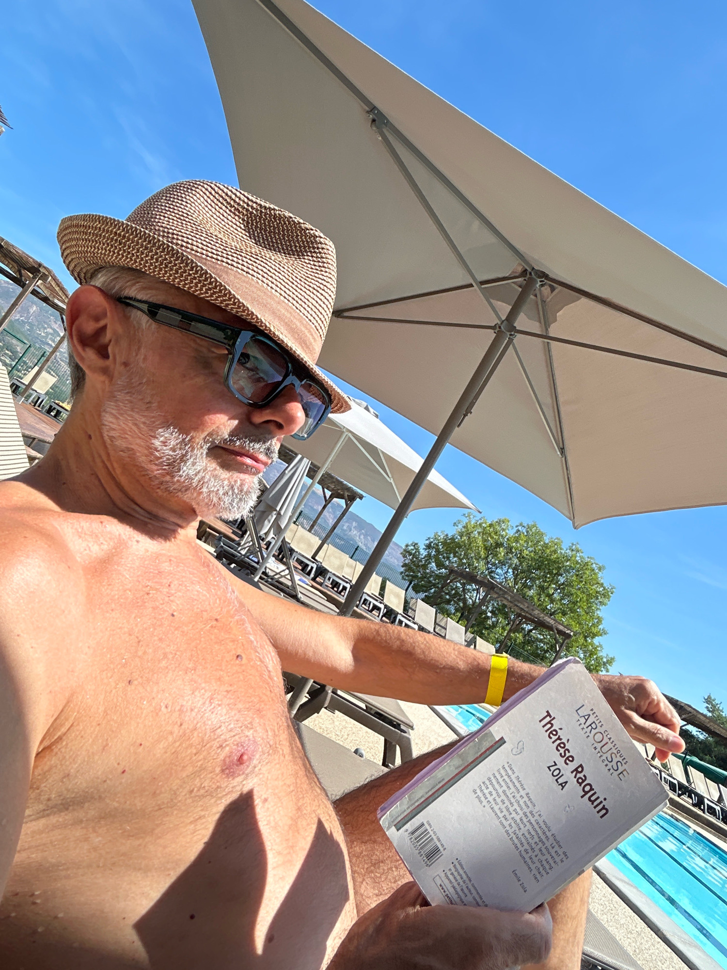 In Le Poët-Célard, France, a man with a beard and wearing a hat and sunglasses is reading a book by the pool. The man is shirtless. He is sitting on a lounge chair under a large umbrella. The water in the pool is blue. There are trees and mountains in the background. The sky is clear and blue.
