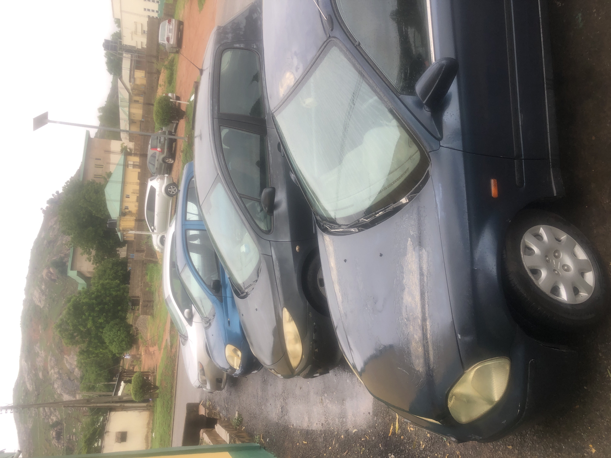 In Jalingo, Nigeria, a line of parked cars is visible on a wet, asphalt road. The cars, from front to back, include a dark blue sedan with a yellowed headlight and a silver hubcap, a dark gray hatchback with tinted windows and a visible side mirror, a blue sedan with its hood and headlight visible, and a silver hatchback. The scene is set against a backdrop of buildings with green and yellow roofs, a paved area, and a rocky, tree-covered hillside. A utility pole with a solar panel stands near the cars. The ground appears to be damp, suggesting recent rain. No people are clearly visible.