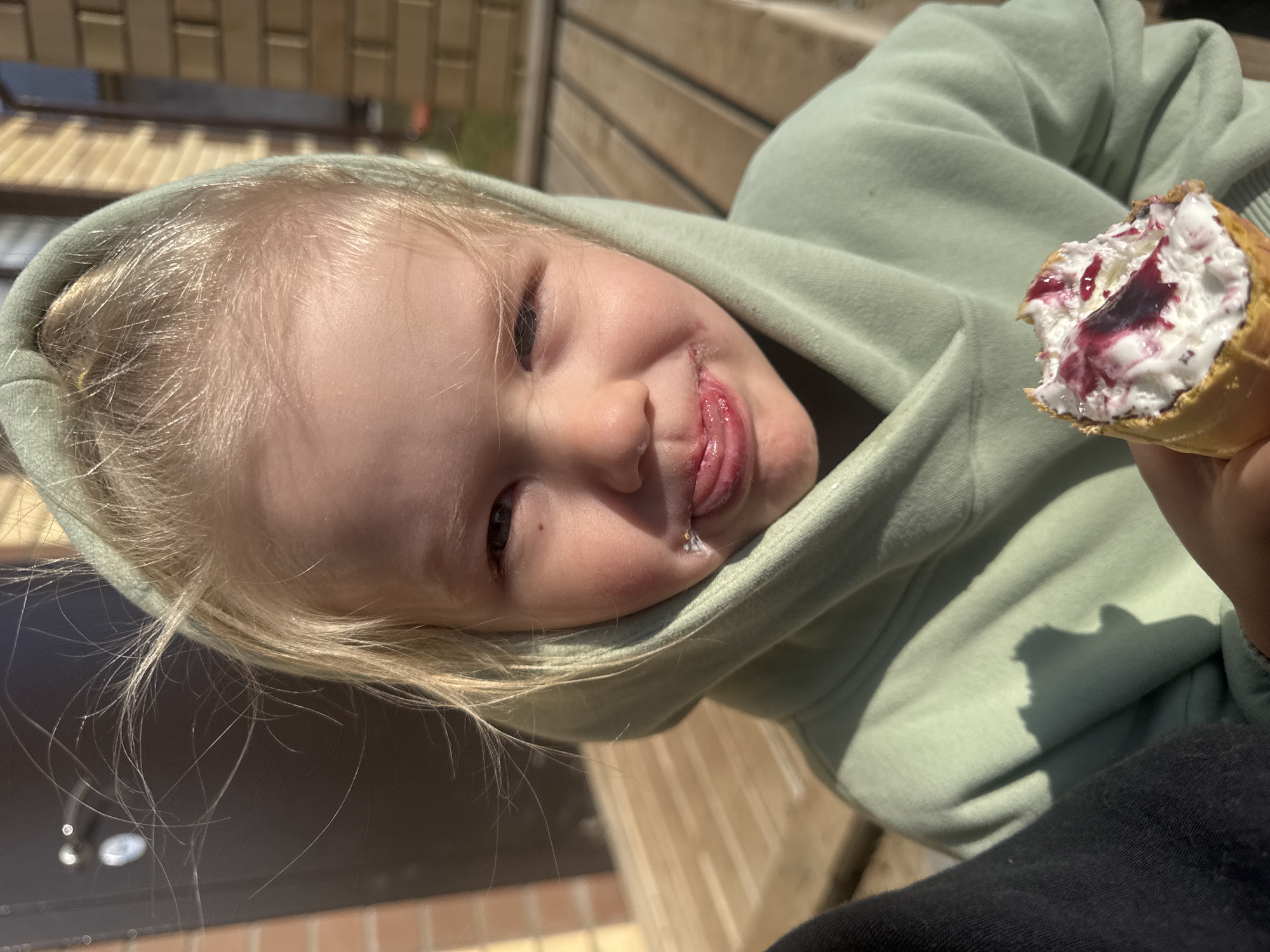 A young, fair-skinned child with light blonde hair is wearing a green hooded sweatshirt. The child is holding a partially eaten ice cream cone, filled with white cream and a dark red topping, likely fruit preserves. The child is sticking their tongue out, with a small drop of saliva visible. The location appears to be outdoors, possibly near a building with a wooden facade. The image was taken in Gornyi, Dvurechenskoe S/P, Russia.