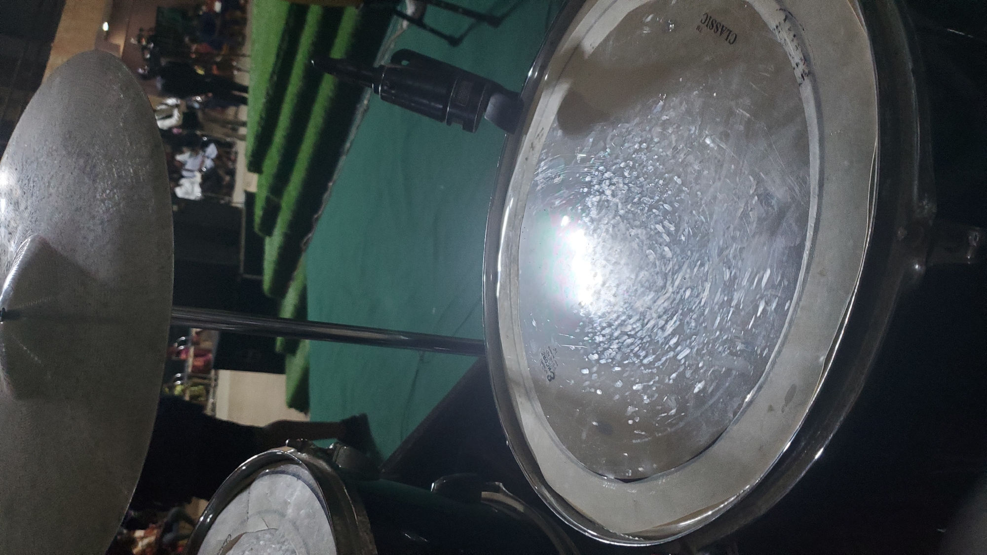 The image shows a close-up view of a drum set, with a cymbal in the upper portion and a drum head in the foreground. The drum head has a reflective surface with visible scratches and is labeled "CLASSIC". A microphone is attached to the rim of the drum. In the background, there are people and tiered green platforms, suggestive of an indoor or outdoor event space. The overall impression is of a performance or rehearsal setting. The location is Jalingo, Nigeria, implied by the context provided in the prompt.