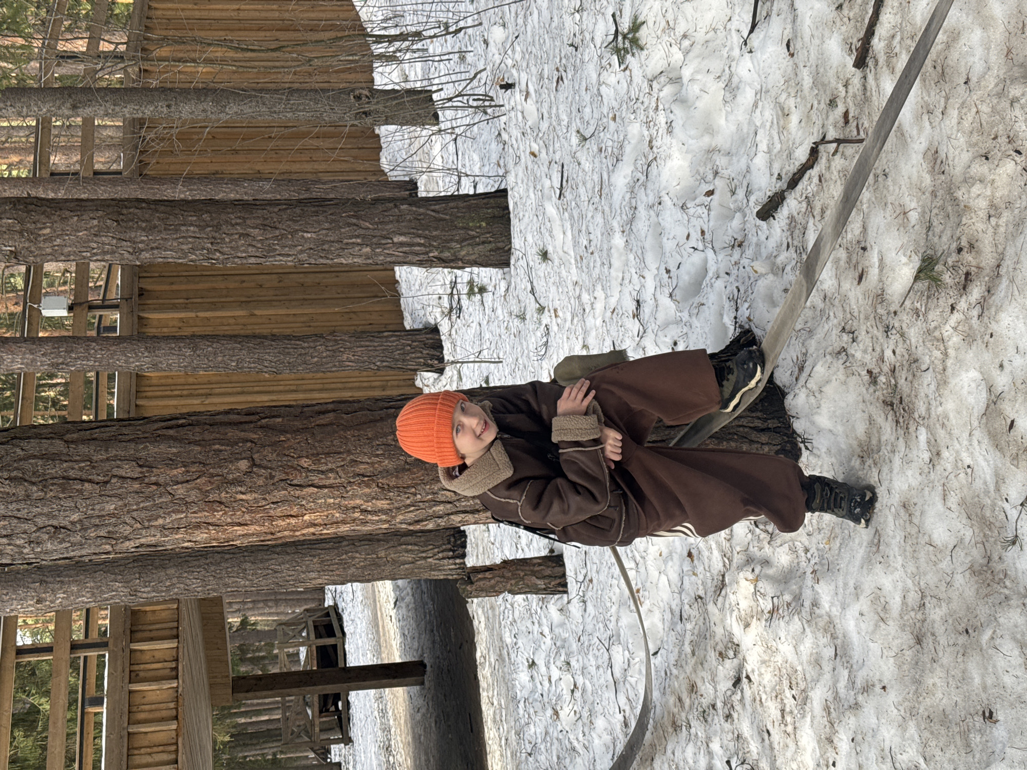 A person wearing an orange beanie, brown coat with fur trim, and brown pants is seated on a metal swing in a snowy environment. The background includes trees and wooden structures that suggest a park or outdoor recreational area, possibly located in or near the city of Votkinsk, Russia, based on provided location context. The scene is likely captured during winter due to the presence of snow on the ground.