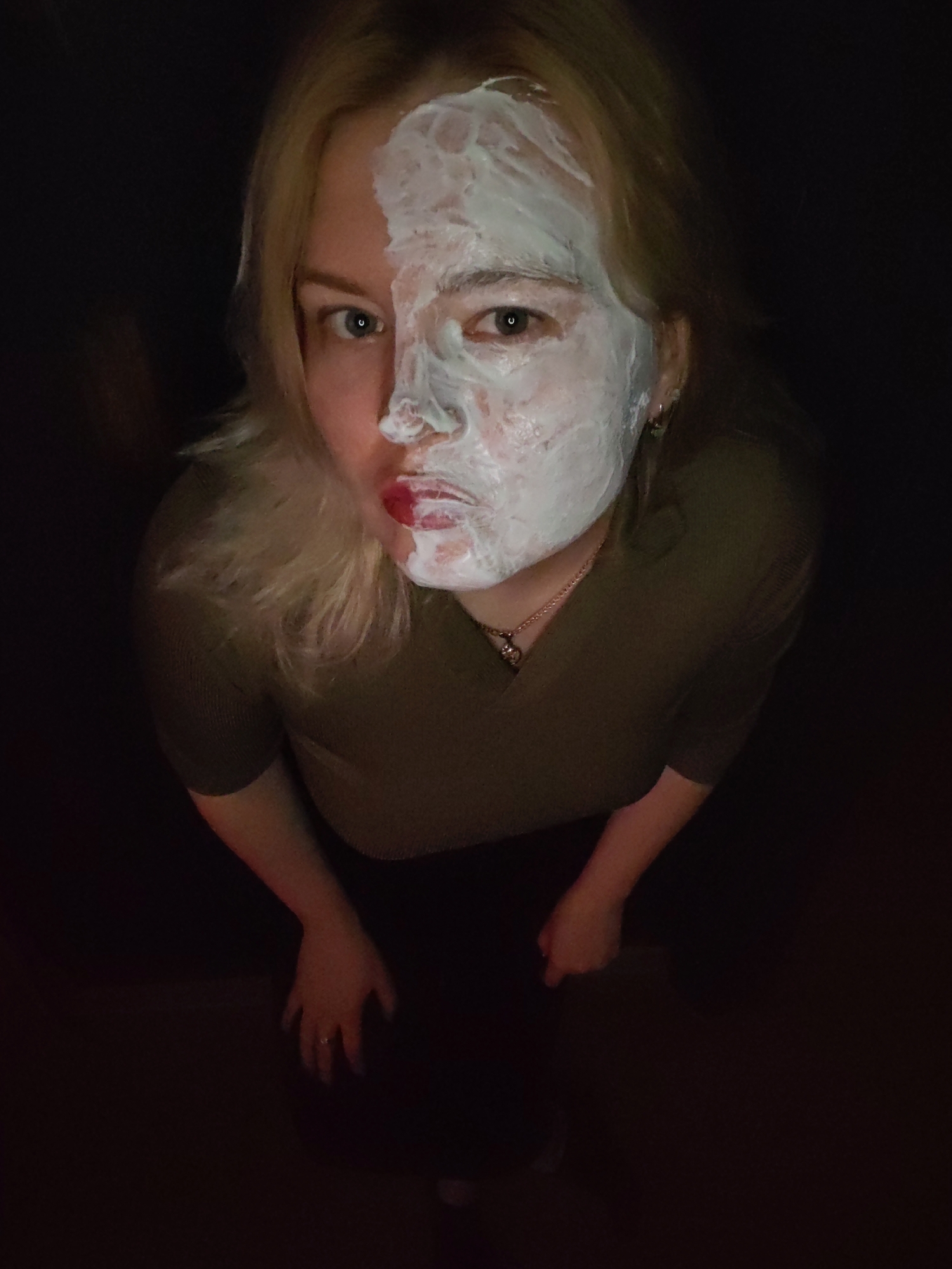 An overhead shot captures a blonde woman in a dimly lit, dark environment, looking directly at the camera. The woman, possibly in her 20s or 30s, is wearing a dark olive or green long-sleeved ribbed top. A golden chain necklace with a small pendant is visible around her neck. A silver ring adorns her left ring finger.
The most prominent feature is her face, which is partially covered by a thick, white or light green creamy face mask. The mask covers the right half of her face, including her forehead, cheek, nose, and upper lip, creating a strong contrast with her unmasked left side. Her left eye is clearly visible, blue or green in color, gazing directly forward. Her right eye is mostly obscured by the mask. Her lips are painted a vibrant red, but some of the face mask has smudged onto her upper lip, mixing with the lipstick.
The lighting appears to come from directly above or slightly in front of her, illuminating her face and casting the background into deep shadow. The perspective is from below, looking up, which emphasizes her face. The activity depicted is likely a beauty or skincare routine. No text is visible in the image. The provided location context is Izhevsk, Russia.