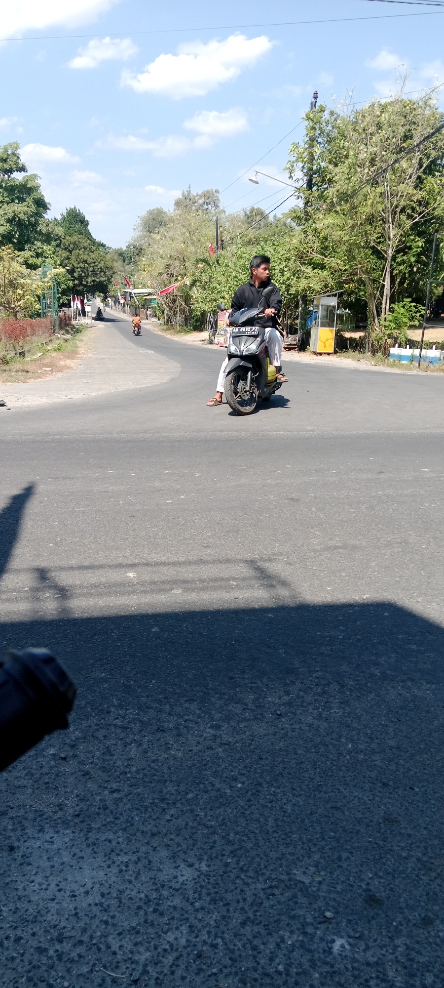 A young man is riding a dark gray scooter on an asphalt road in Maguwoharjo, Indonesia. He is wearing a black long-sleeved shirt, white pants, and brown sandals. The scooter has a license plate that reads "AB 6852 PJ". The road is a two-lane asphalt road with a white dashed line marking the lanes. On the left side of the road, there is a sidewalk with trees and foliage. On the right side of the road, there is a yellow and white kiosk and more trees. The sky is blue with white clouds, and the sun is shining. There is another scooter in the distance on the road, also with a rider. The perspective of the image is from a low angle, with shadows of objects on the left and right of the frame cast across the road.