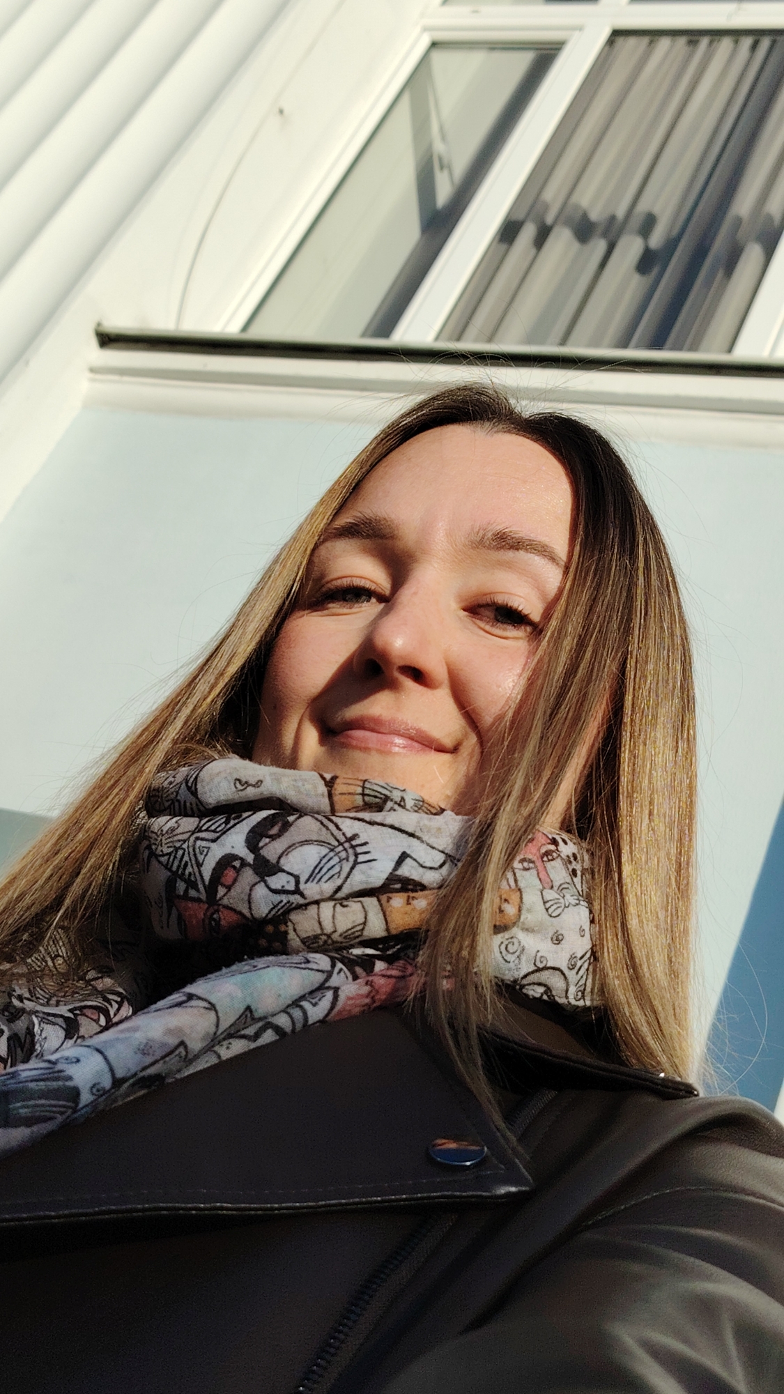 A low-angle close-up portrait captures a young woman smiling directly at the camera, suggesting a selfie. She has long, straight, light brown hair with blonde highlights, which flows around her shoulders. Her face is brightly illuminated by direct sunlight, casting some shadows and highlighting her cheerful expression. She is wearing a dark, possibly black, leather-style jacket with a visible zipper and a silver-toned button on the collar. Around her neck, she wears a light-colored scarf patterned with whimsical, cartoon-like illustrations of animals or figures outlined in black, with soft pastel color accents. The background features the exterior of a modern building, characterized by white-framed windows and light blue or white walls. Reflections in the windows indicate an interior with curtains. The strong sunlight suggests it is a clear day, likely morning or afternoon. No visible text is present.