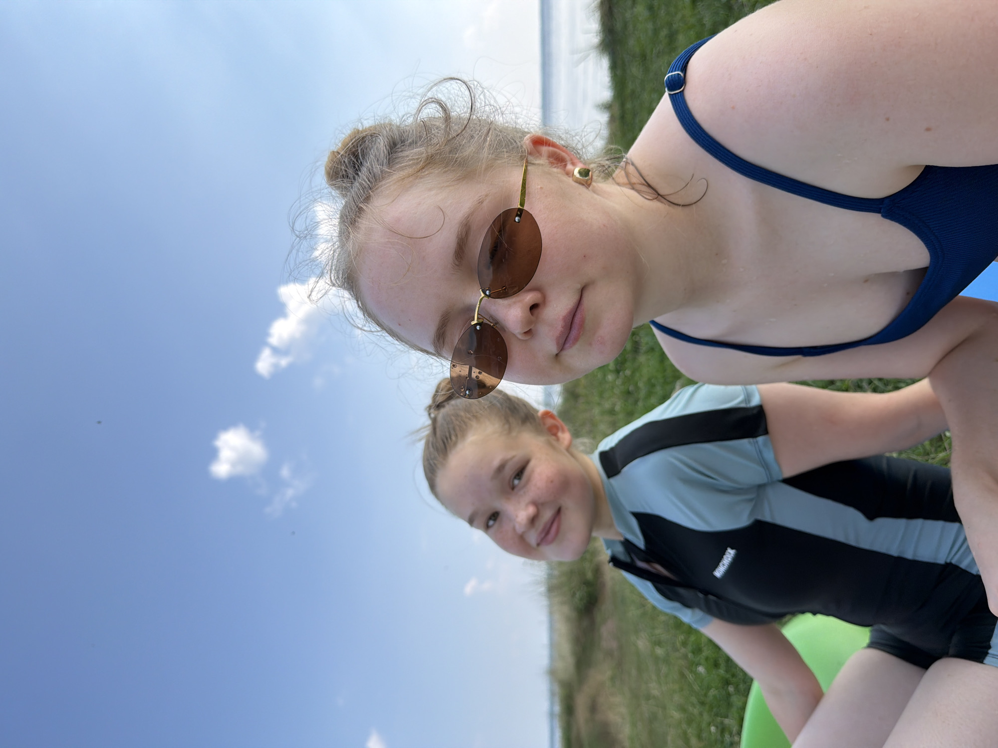 Two young females are sitting outdoors, facing the camera. The female on the left is wearing a black and light blue rash guard with a zipper in the front and the text "MONCHOIX" visible on it. She has light skin, freckles, and her hair is pulled back in a ponytail. She is looking towards the camera with a slight smile. The female on the right is wearing a dark blue swimsuit. She has light skin, her hair is in a bun, and she is wearing oval-shaped brown sunglasses and a gold earring in her right ear. She is looking directly at the camera with a neutral expression.
They are sitting on what appears to be a green inflatable object. The background consists of a grassy area with some small white flowers. Beyond the grass, there is a body of water, possibly a lake or river, and a hazy shoreline with some vegetation. The sky is blue with scattered white clouds.
The location is not specified in the image or the prompt.