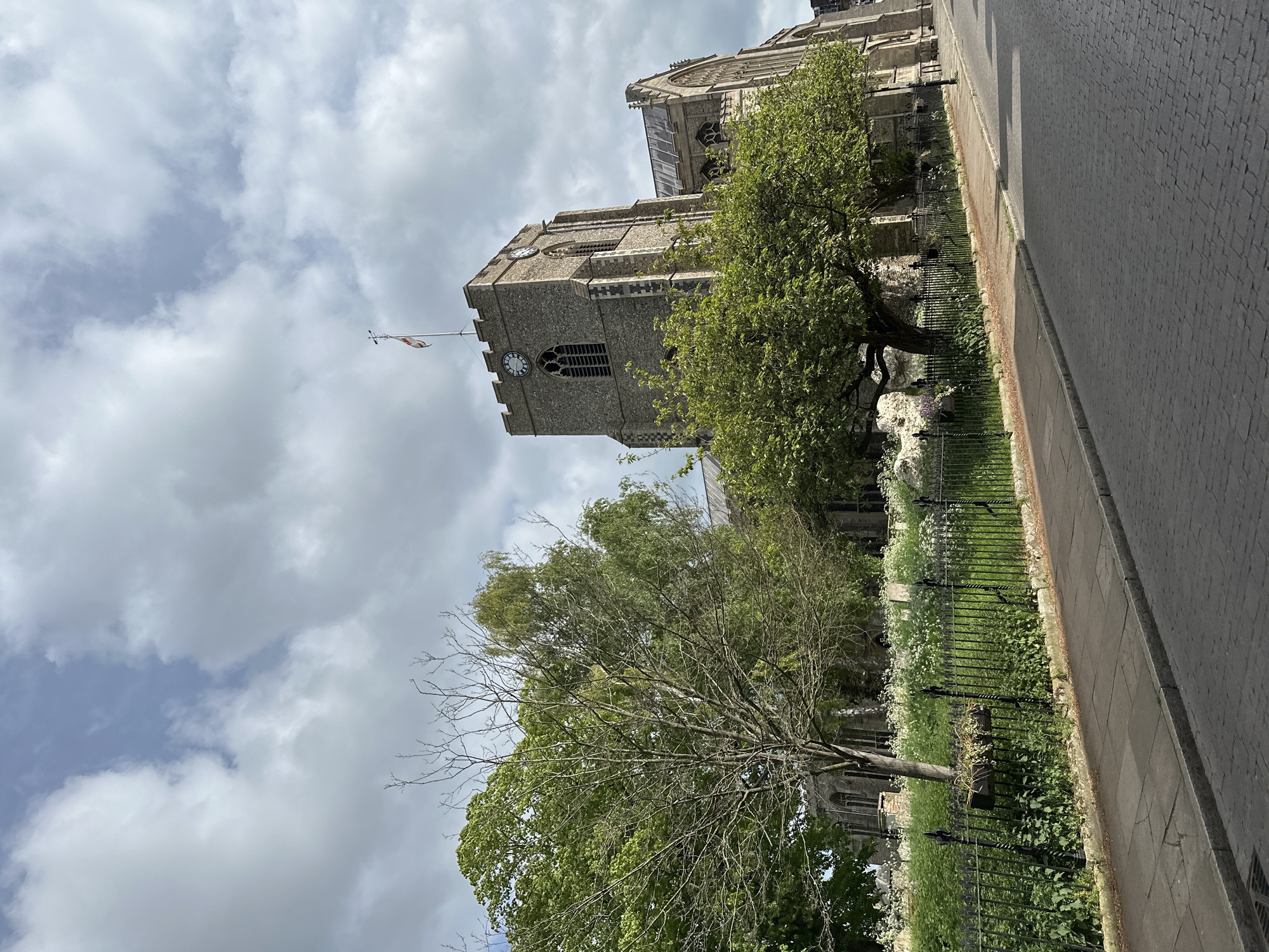 The image shows a building, likely a church, in West Suffolk, United Kingdom. The building is constructed of grey stone and features a tower with a clock face. A flag is flying from the top of the tower. The church is surrounded by trees and a black metal fence. The street is paved with cobblestones. The sky is overcast with grey clouds.