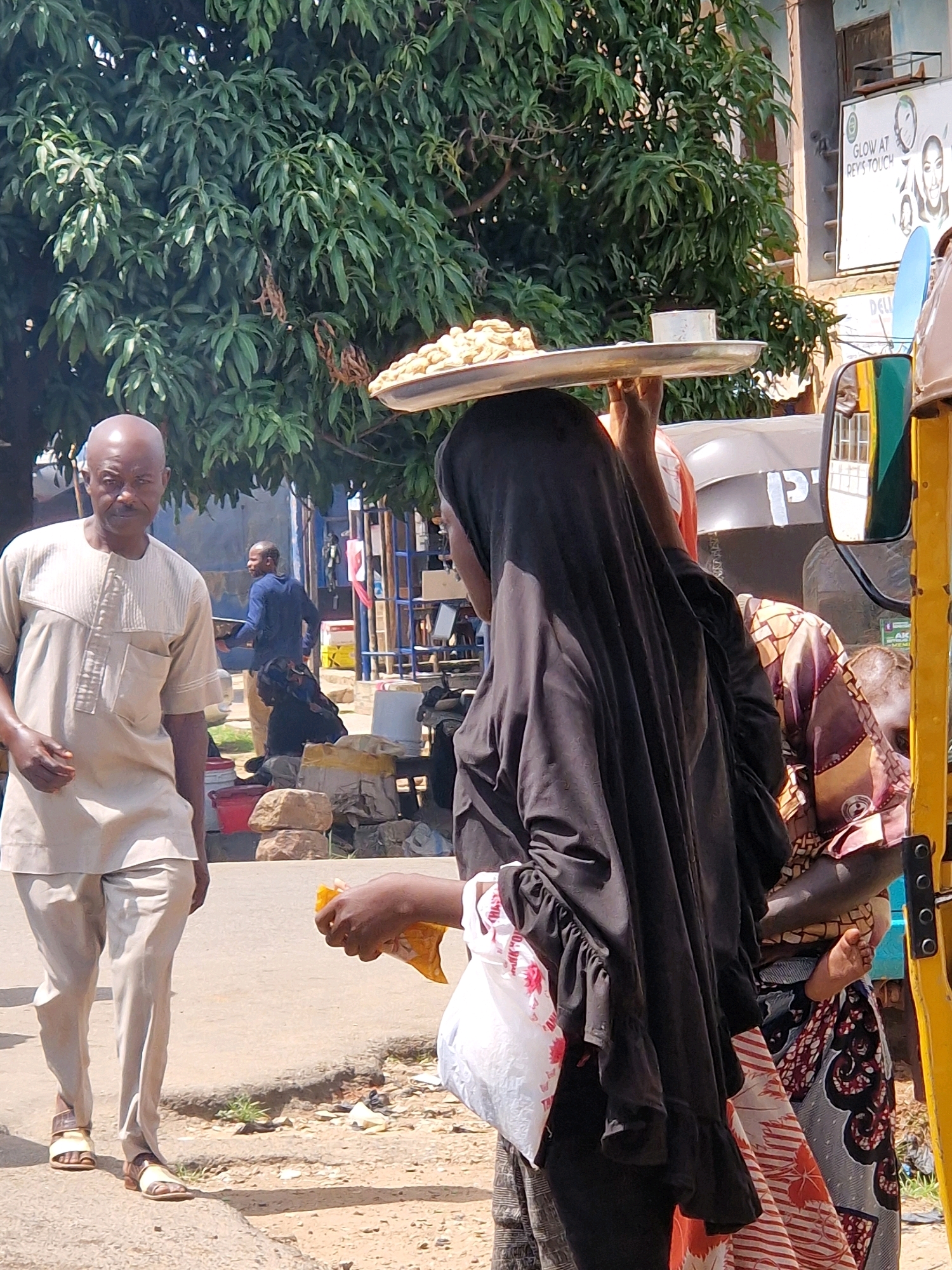 In Jos, Nigeria, an outdoor market scene unfolds. A bald Black man, wearing a light-colored traditional outfit, stands facing left, his gaze directed forward. To his right, a Black woman, veiled in a black hijab, carries a large metal tray laden with small baked goods balanced on her head. She is seen from the side, her left hand reaching towards her chest, clutching a plastic bag. Behind her, another Black woman in a colorful patterned fabric dress and headscarf is partially visible, her arm extended. In the background, a man in a blue shirt walks away, and elements of market stalls, including red and yellow structures, are present. The ground is paved with concrete and dirt, with scattered stones and debris. Lush green trees provide shade over the scene. A portion of a yellow vehicle, likely a tuk-tuk, is visible on the far right.