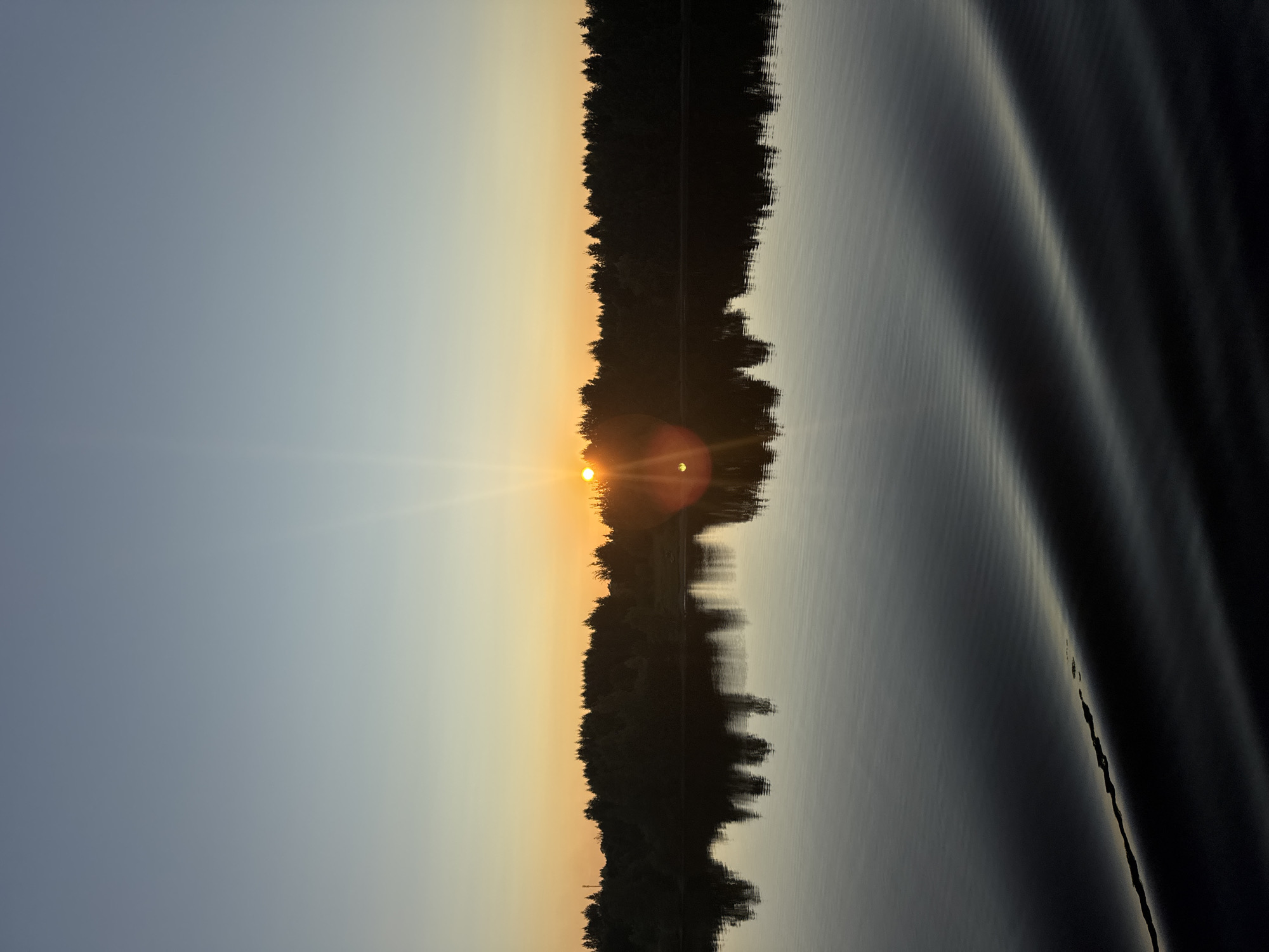 A serene sunset over a calm lake is depicted in this vertical, eye-level shot. The sun, a bright orb, is low on the horizon, casting a warm orange glow that transitions to a soft yellow and then a pale blue in the sky. Rays of light emanate from the sun, creating a lens flare effect. A dense forest lines the far bank of the lake, silhouetted against the vibrant sky. The trees' reflections are clearly mirrored on the glassy surface of the water. In the foreground, gentle ripples disturb the water's surface, catching the fading light. The scene evokes a sense of tranquility and natural beauty. The location is identified as Nova, Russia, implying it is a natural setting within or near the city. There are no people or man-made structures prominently featured in the image, focusing solely on the natural landscape.